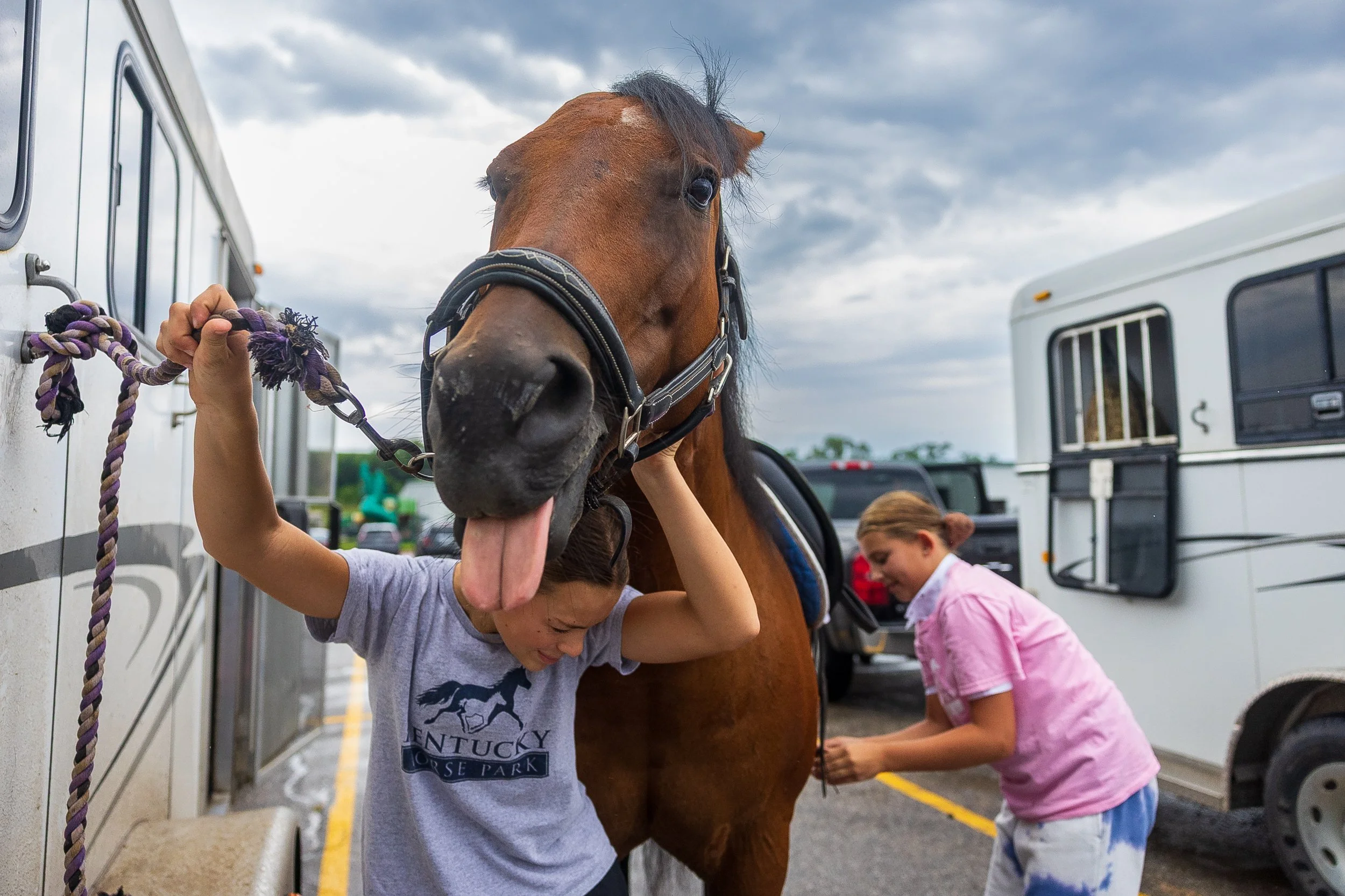 Elsie Maranville ducks underneath her Bay horse Katniss while her friend Saylor Bontrager, right, works to secures her saddle before the Equestrian dressage competition at the Lancaster State Fair on Wednesday, July 29, 2026, at the Sandhills Global 