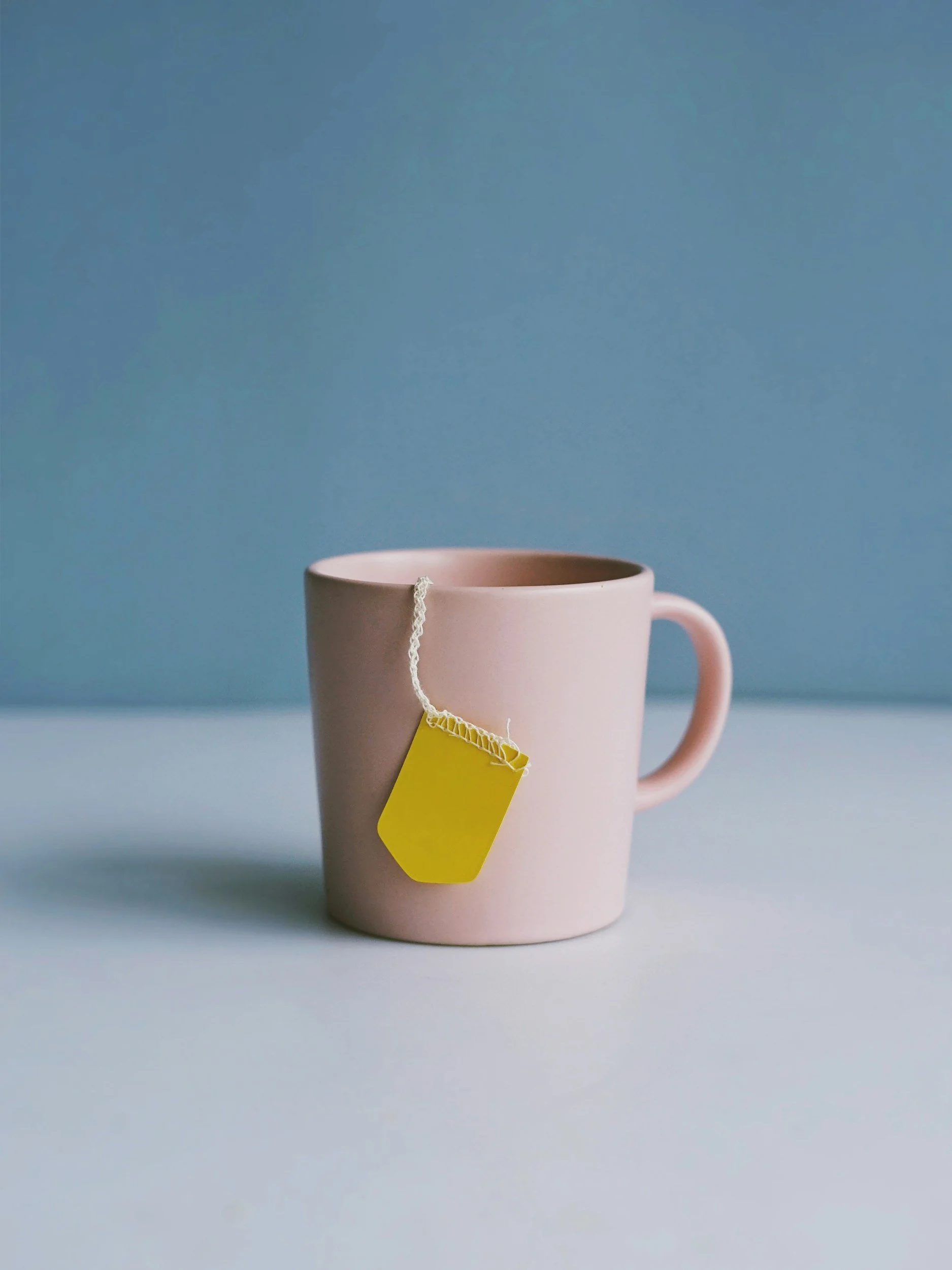 A pink mug with a yellow tag attached to its handle, placed on a light blue surface against a blue background.