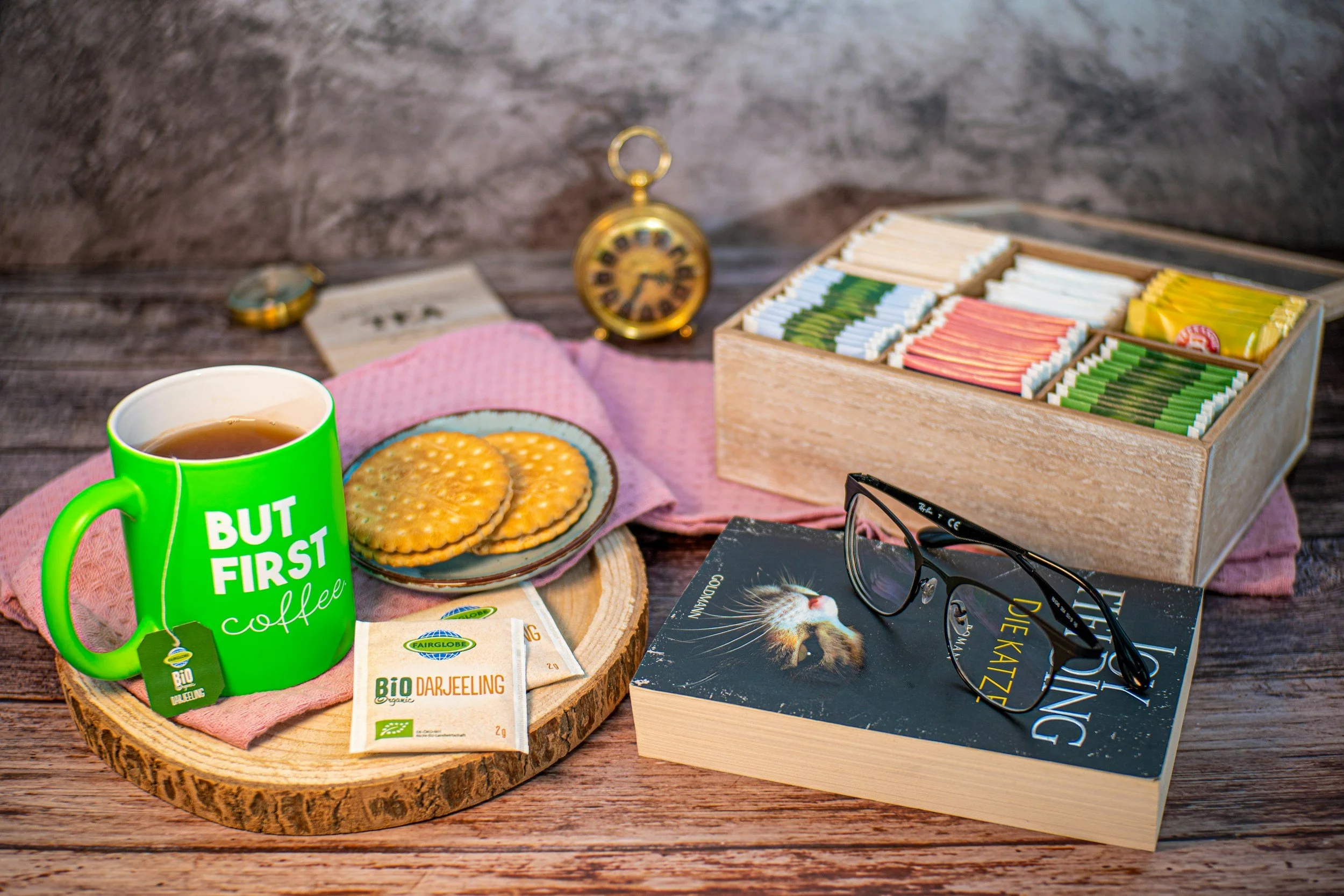 A table with a green mug reading 'But First Coffee,' a plate with biscuits, a box of tea bags, a broken pocket watch, reading glasses, a book, and tea bags on a wooden surface with a textured gray background.