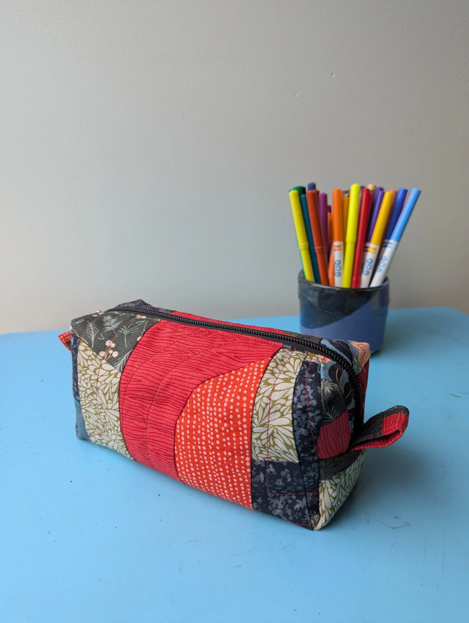 A fabric zippered pouch with a colorful patchwork design and a cluster of multicolored markers inside a black cup on a light blue surface.