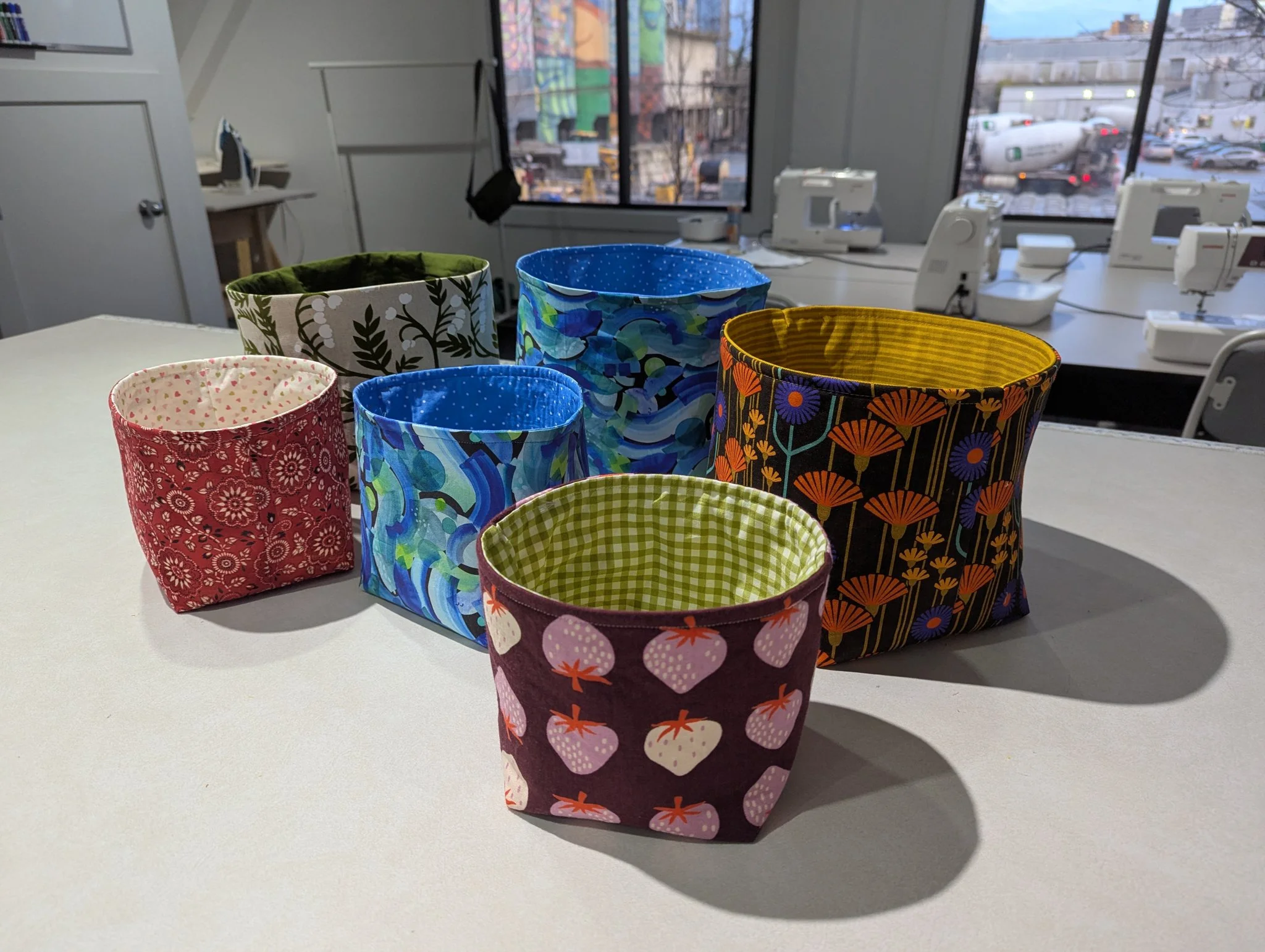 Seven colorful fabric storage bins of various patterns and sizes are displayed on a white table. The bins include floral, geometric, and patterned prints in red, green, blue, purple, and black. The background shows a sewing workshop with sewing machines, tables, chairs, and large windows revealing parked cars outside.