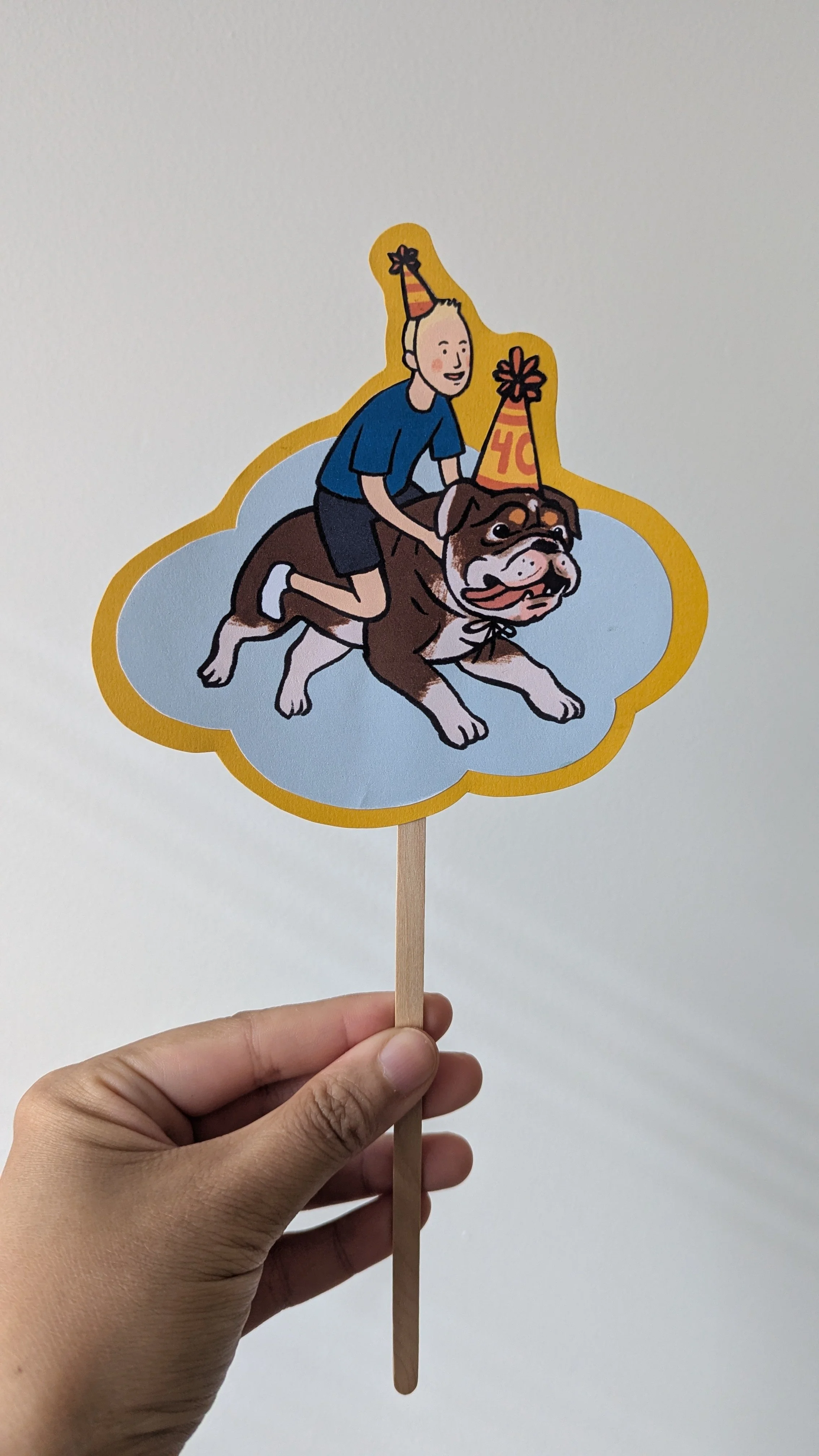 Hand holding a paper cutout of a boy riding a large dog, both wearing birthday hats, on a stick.