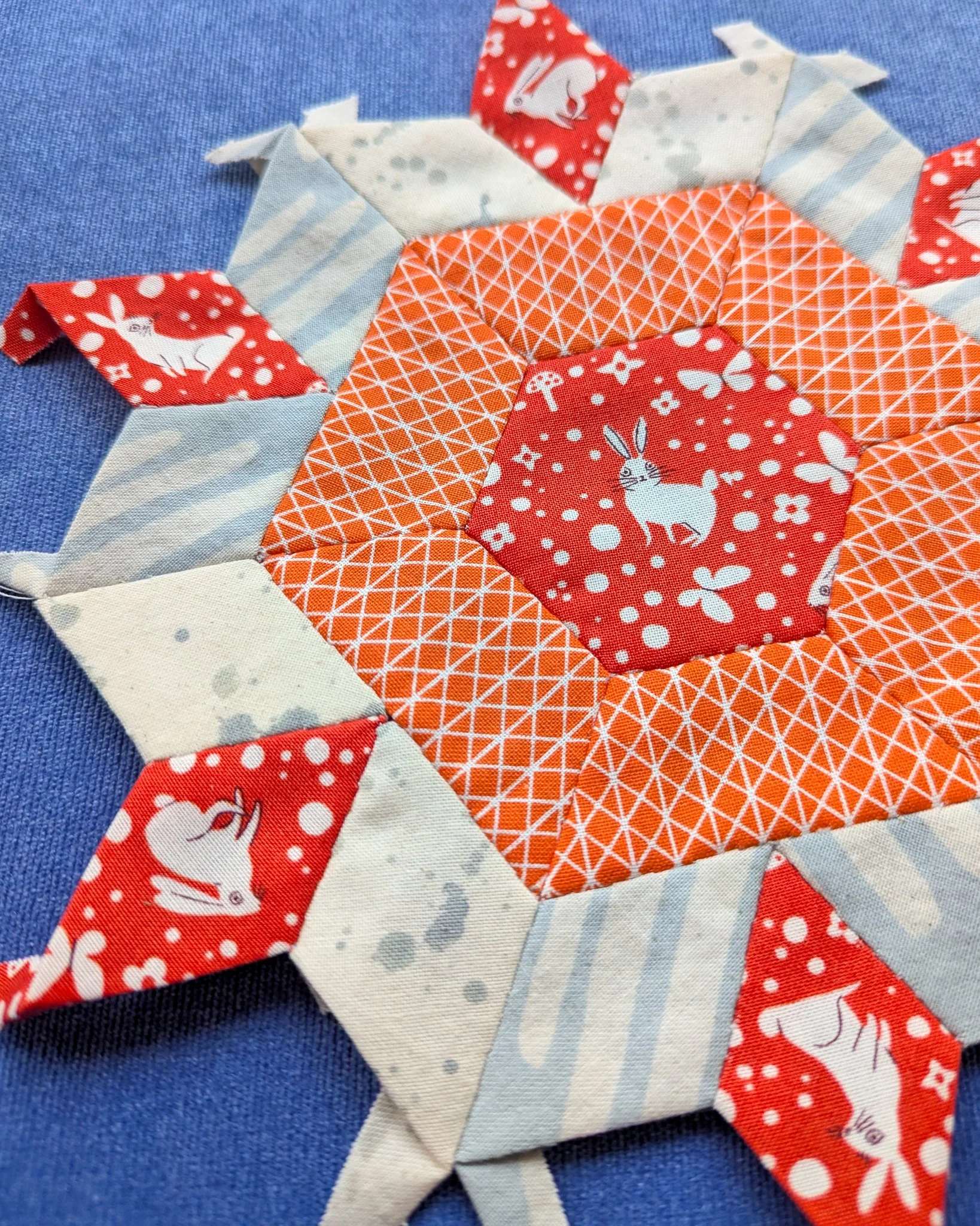 Close-up of a quilted fabric pattern with a central red circle featuring a white rabbit, surrounded by orange, white, and red fabric pieces with bunny, butterfly, and floral designs, on a blue fabric background.