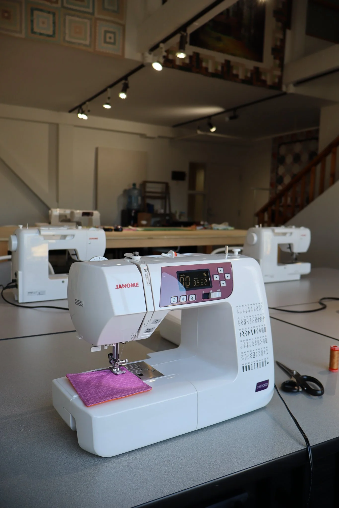 A sewing machine on a table, with additional sewing machines in the background, in a sewing studio decorated with quilted hangings