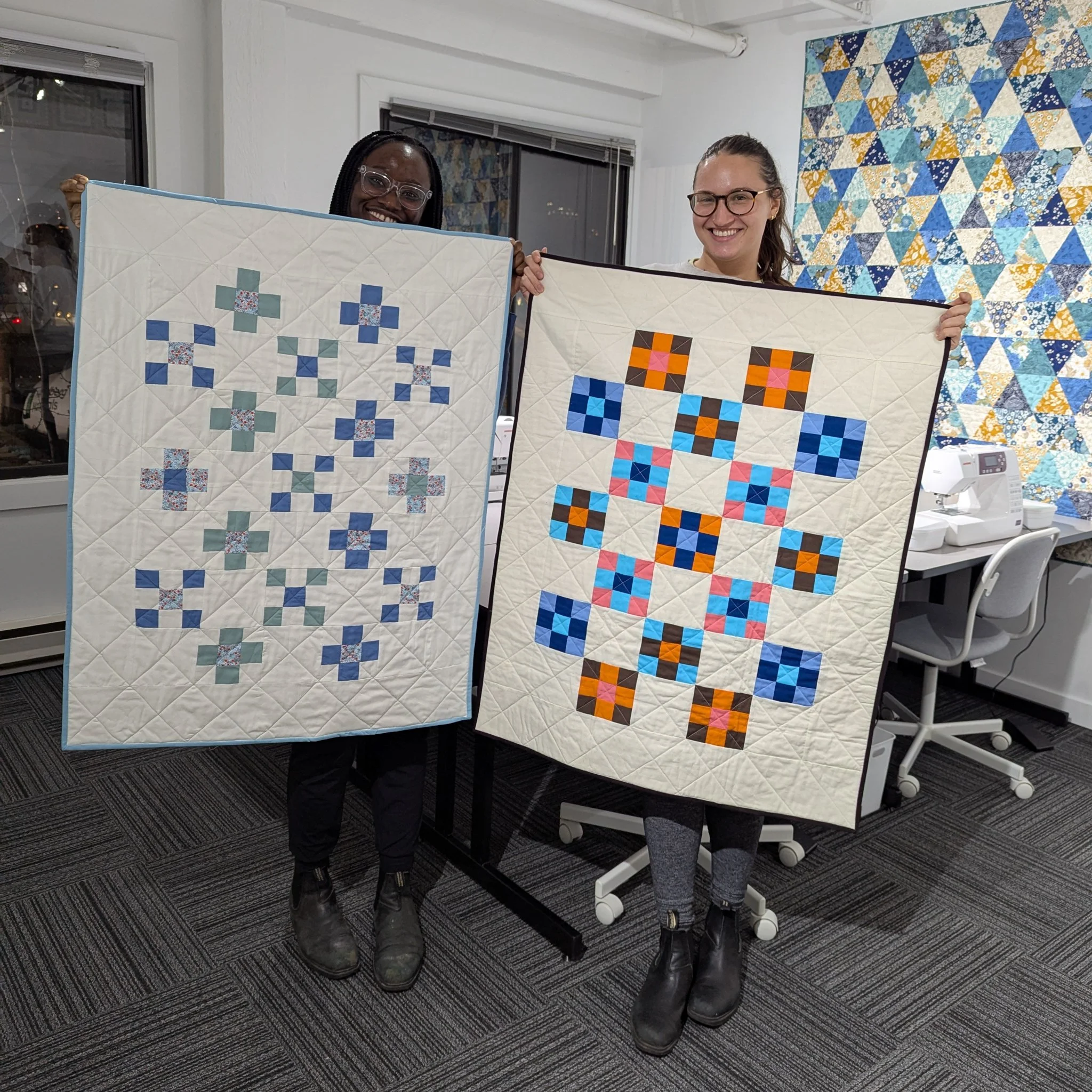 Two women holding up completed quilt projects inside a room with sewing machines and a large colorful quilt hanging on the wall.