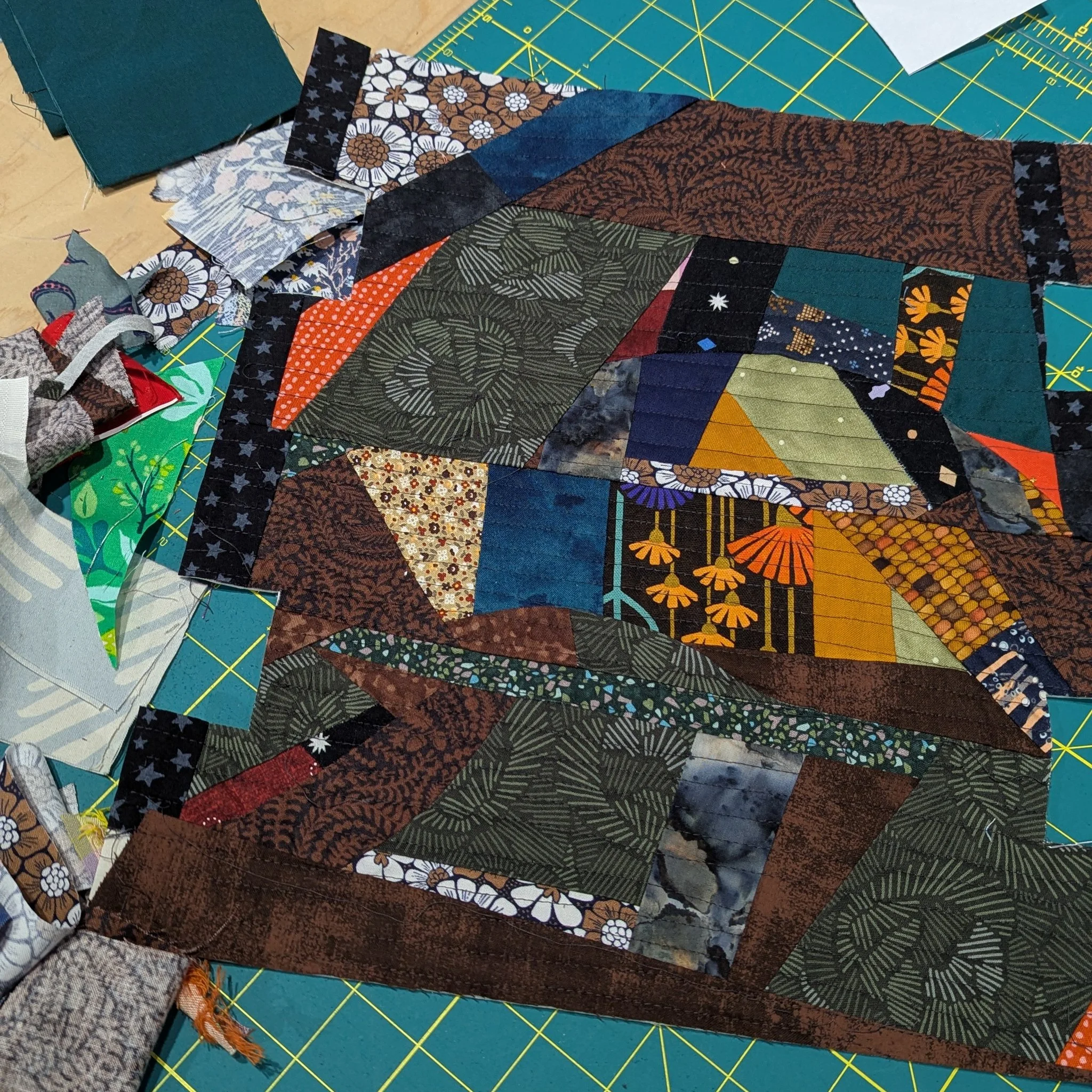Close-up of a colorful patchwork quilt with various fabric patterns, laid out on a cutting mat with grid lines.