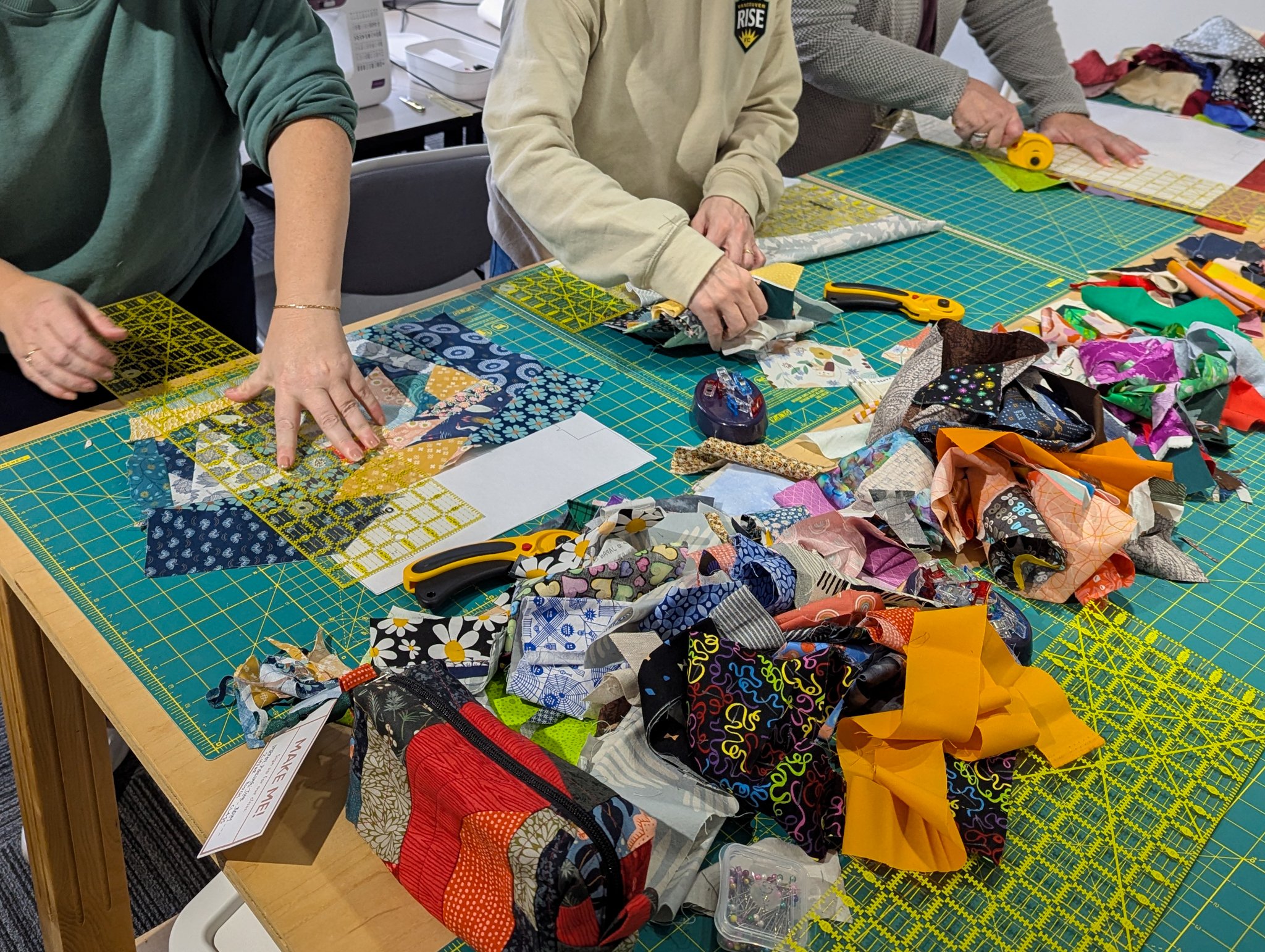 People working on a sewing project with fabric pieces, scissors, and rulers on a blue cutting mat.