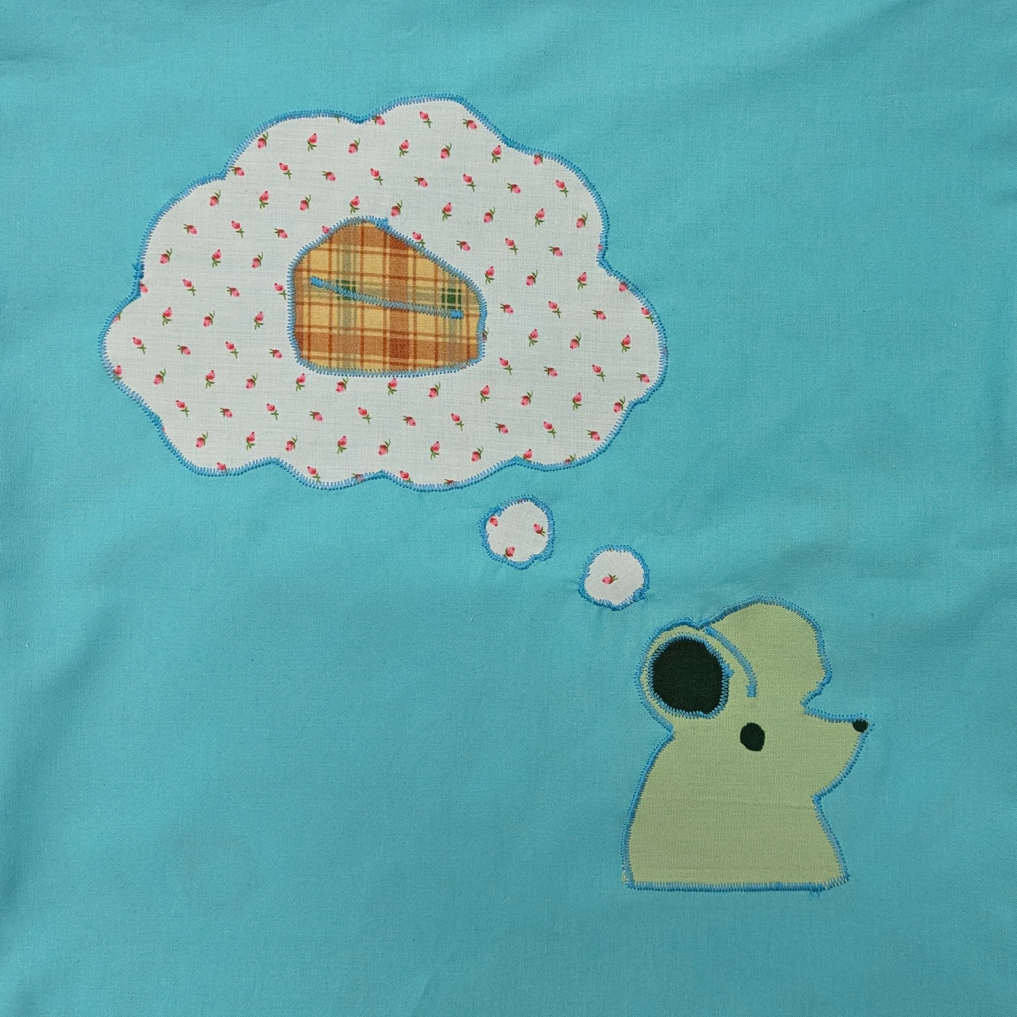 A cute embroidered design of a mouse's head with a thought bubble above it with a wedge of cheese, on a light blue fabric background.