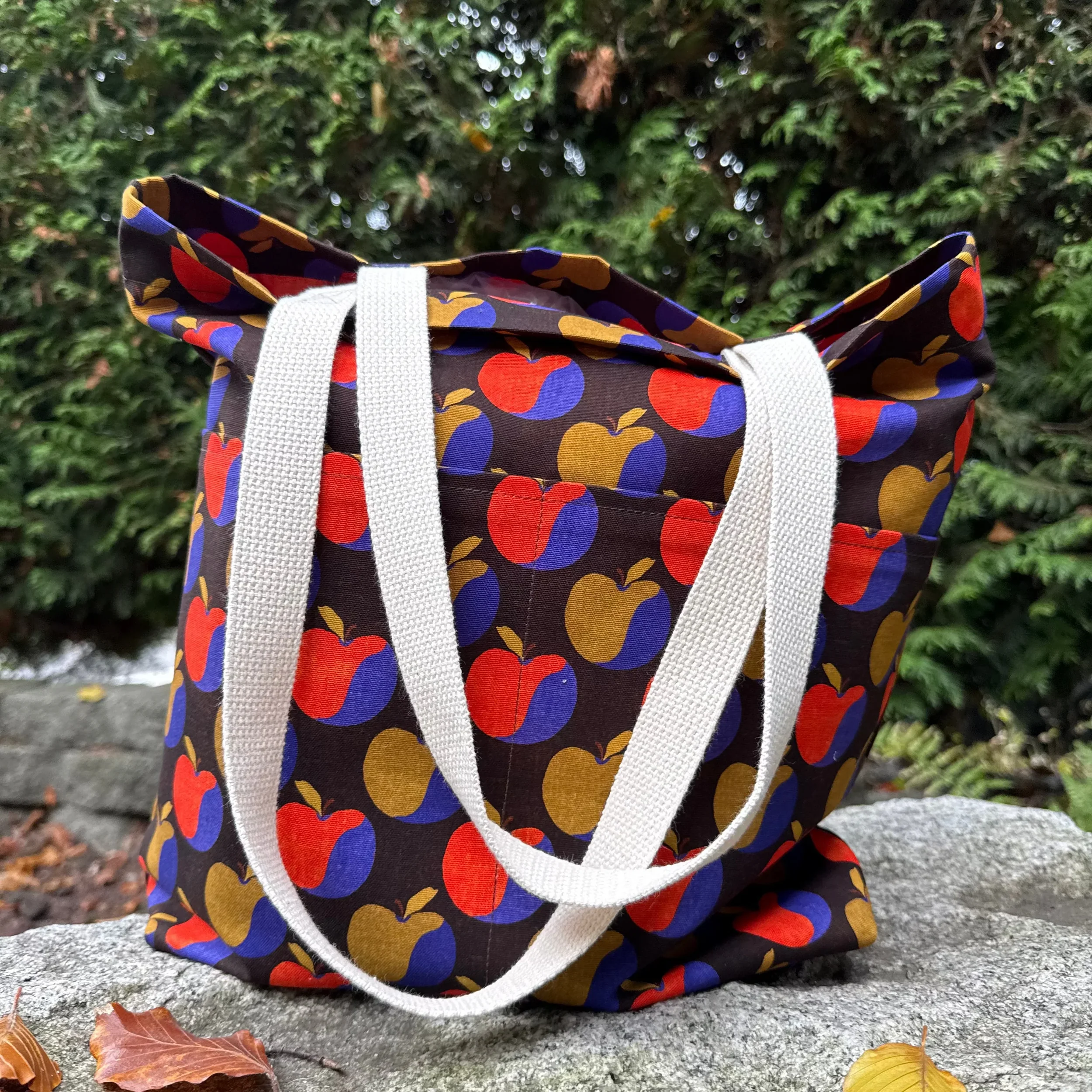 A fabric tote bag with a black background and a colorful apple pattern, placed outdoors on a rock with fallen leaves, in front of green shrubbery.