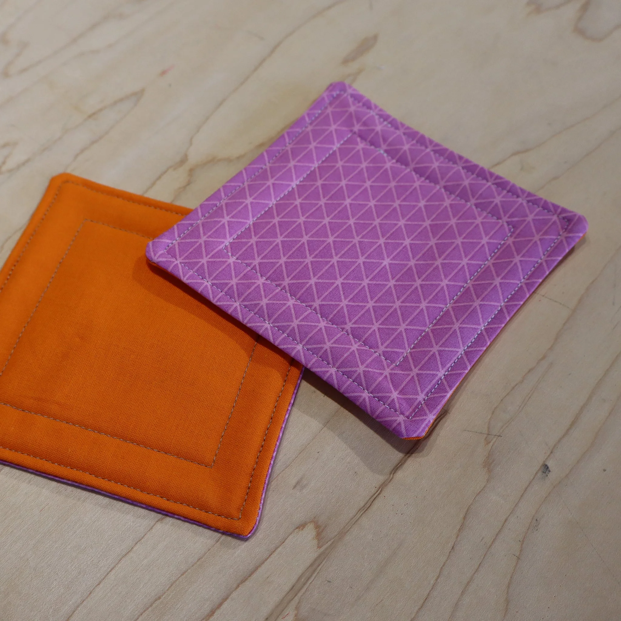 Two colorful fabric coasters, one purple with a geometric pattern and one orange, placed on a wooden surface.