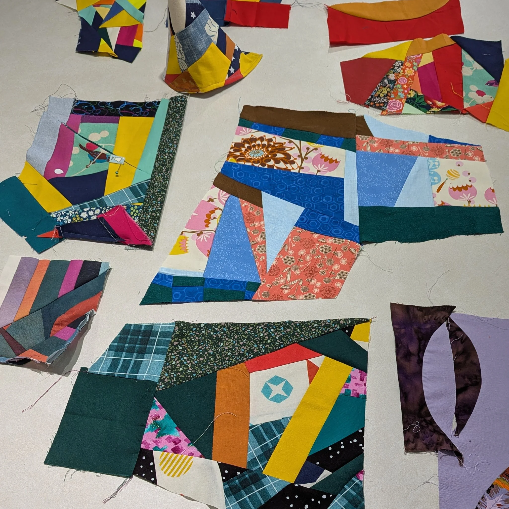 Several quilt blocks created with improv piecing techniques in various colorful patterns and fabrics, some partially sewn or in progress, laid out on a flat surface.