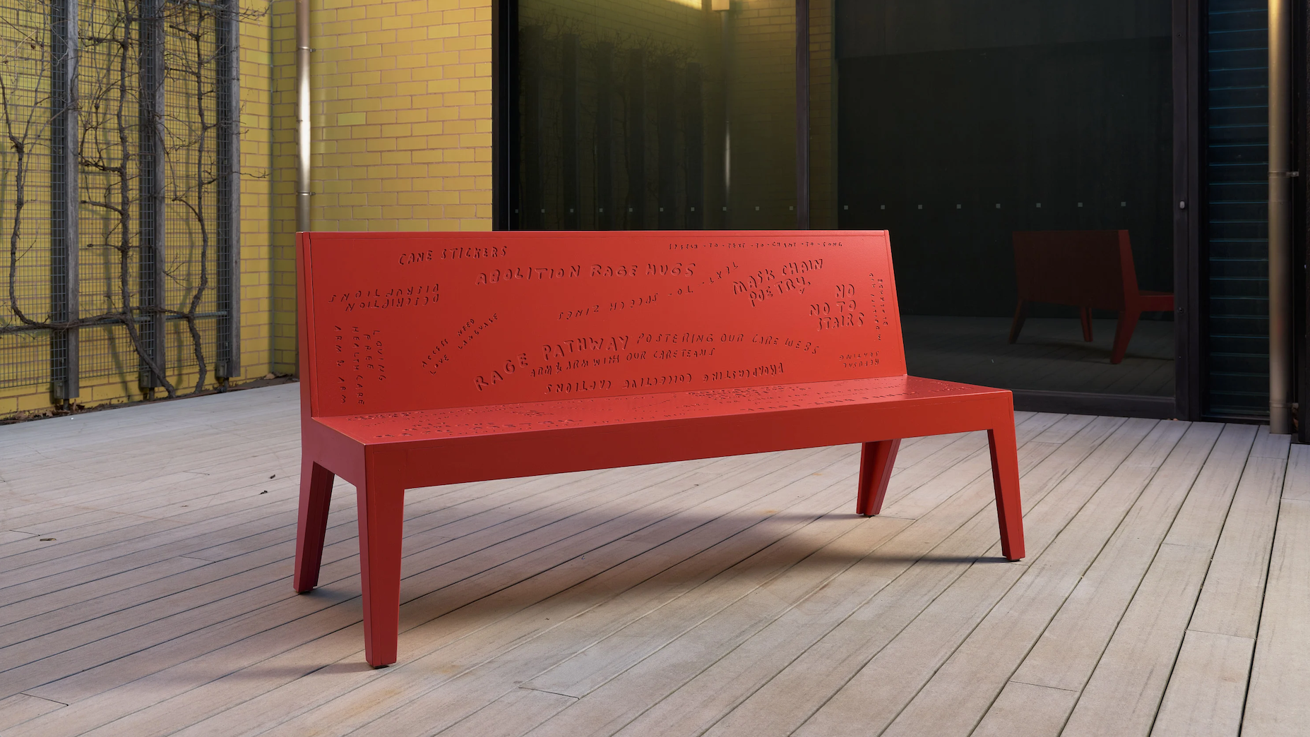 A bench painted in an red-orange color sits in the middle of an outdoor patio, with a yellow wall behind it and a wall with a large window nearby.