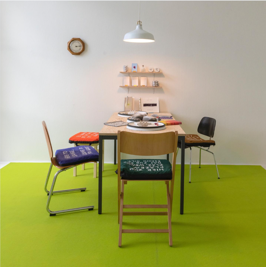 A diningroom-sized-table with chairs on a square of lime green carpet. There are assorted objects on the table and on some small wall shelves.