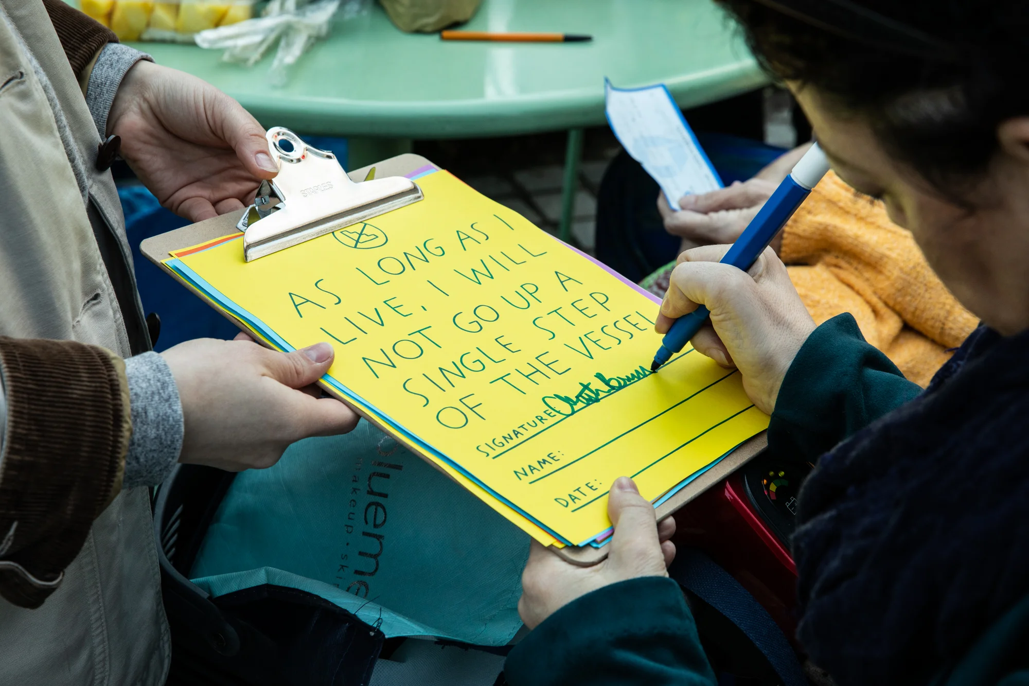 A close-up of Christine Bruno signing their pledge: “As long as I live, I will not go up a single step of the Vessel.” The pledge on colorful paper, riso-printed with blue hand-drawn text, and has a crossed-out-stairs symbol at the top.