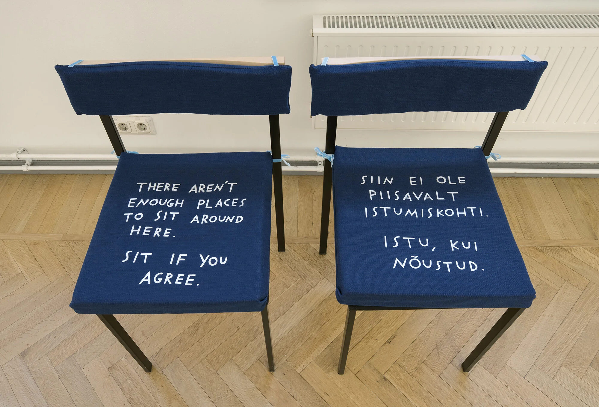 Two chairs with custom blue cushions. Both cushions have white hand-written text. One reads, “There aren’t enough places to sit around here. Sit if you agree.” The other has text in Estonian of a similar length.