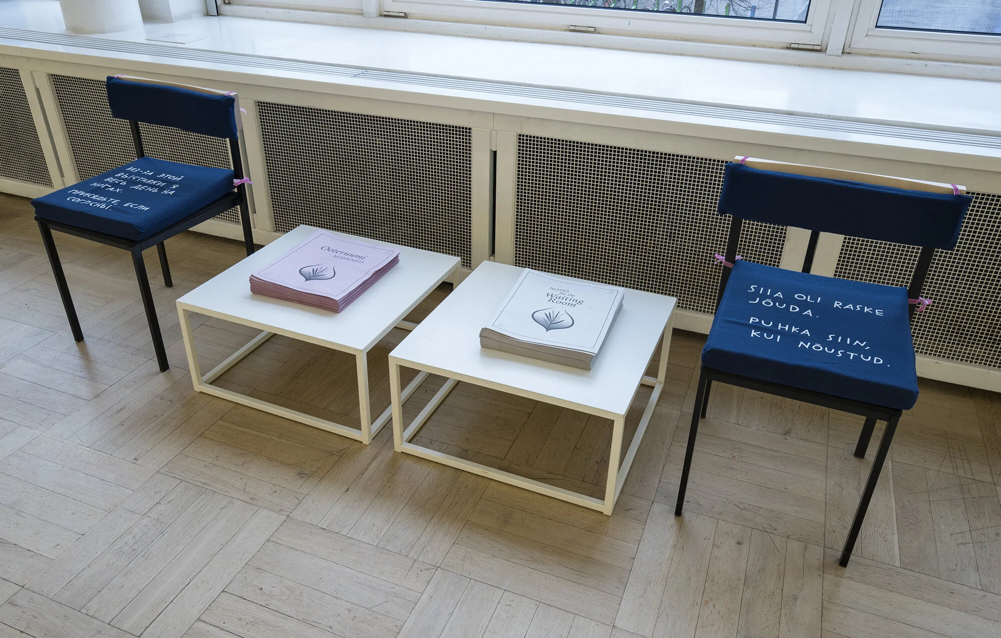 Similar chairs and cushions flanking a table with copies of a broadsheet titled “Notes for the Waiting Room.” One cushion has Russian text, the other Estonian.