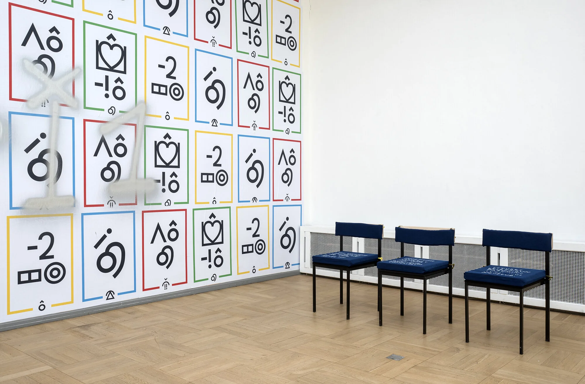 Three similar chairs and cushions line up against a wall. Here the text is not legible because of the angle. The adjacent wall is filled with posters by Jesse Darling featuring Blissymbolics.