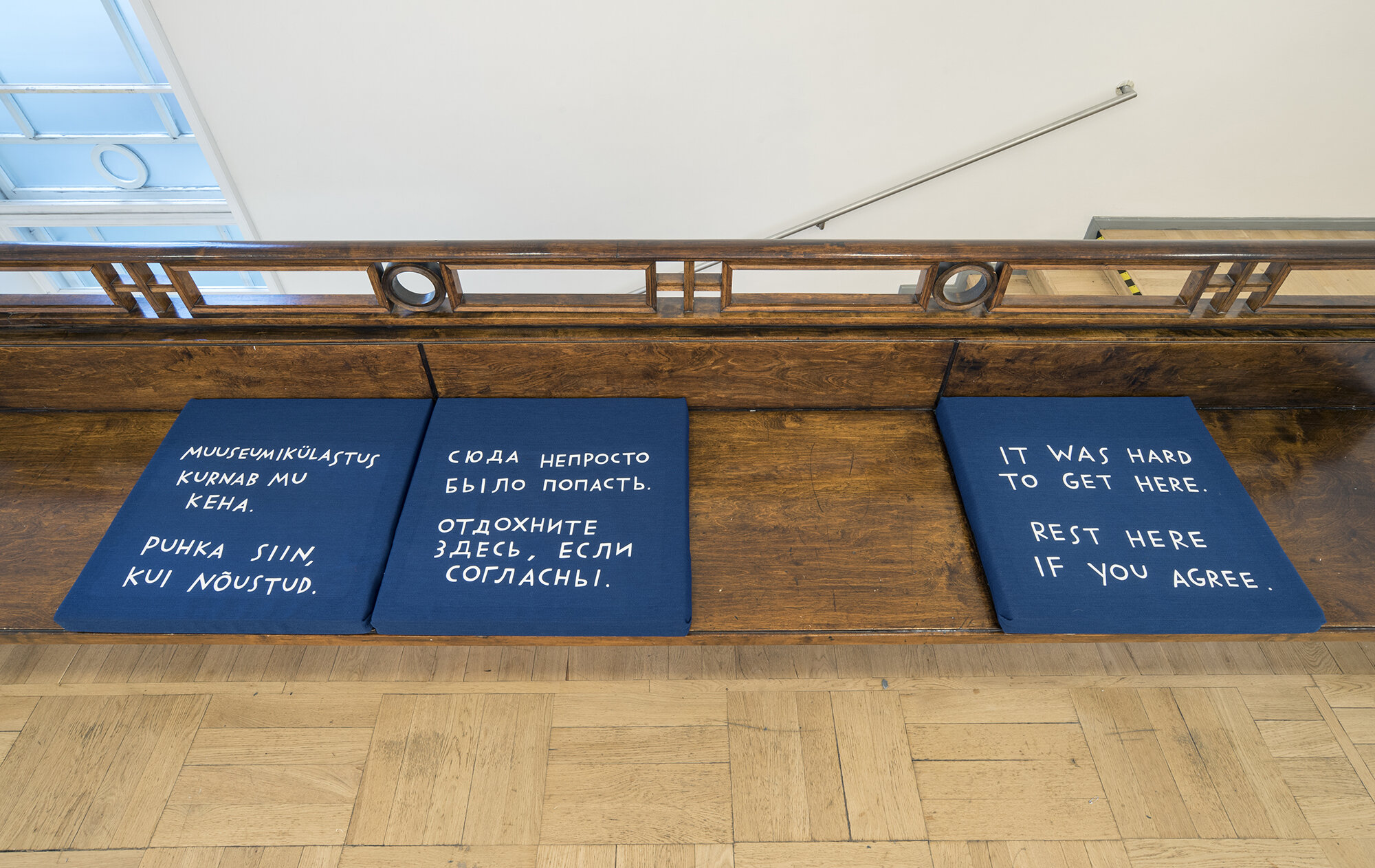 Three cushions fitted to a built-in bench. There is a mix of languages here too. The English-language cushion reads, “It was hard to get here. Rest if you agree.”