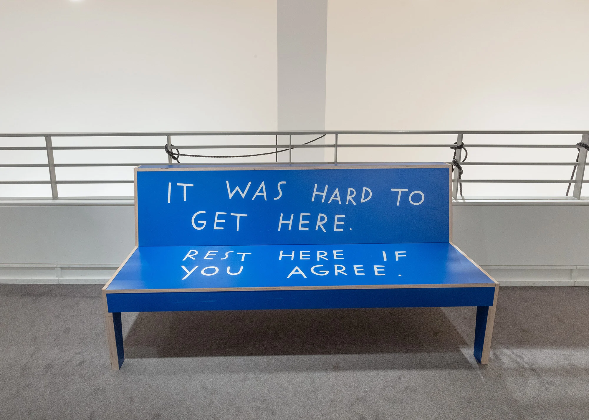 A bright blue bench with white, hand-lettered text that reads, “It was hard to get here. Rest here if you agree.”