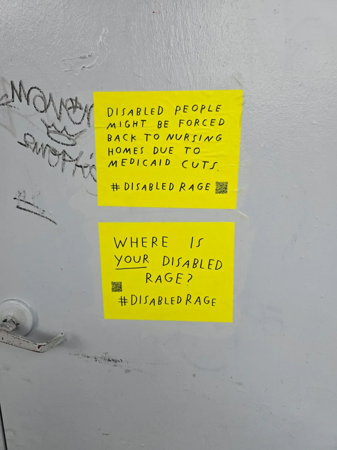 Two signs on top of each other on a gray street door. They are bright yellow with  a QR code and #DisabledRage.  The top sign has a message about Medicaid cuts and one below reads "WHERE IS YOUR DISABLE RAGE?"