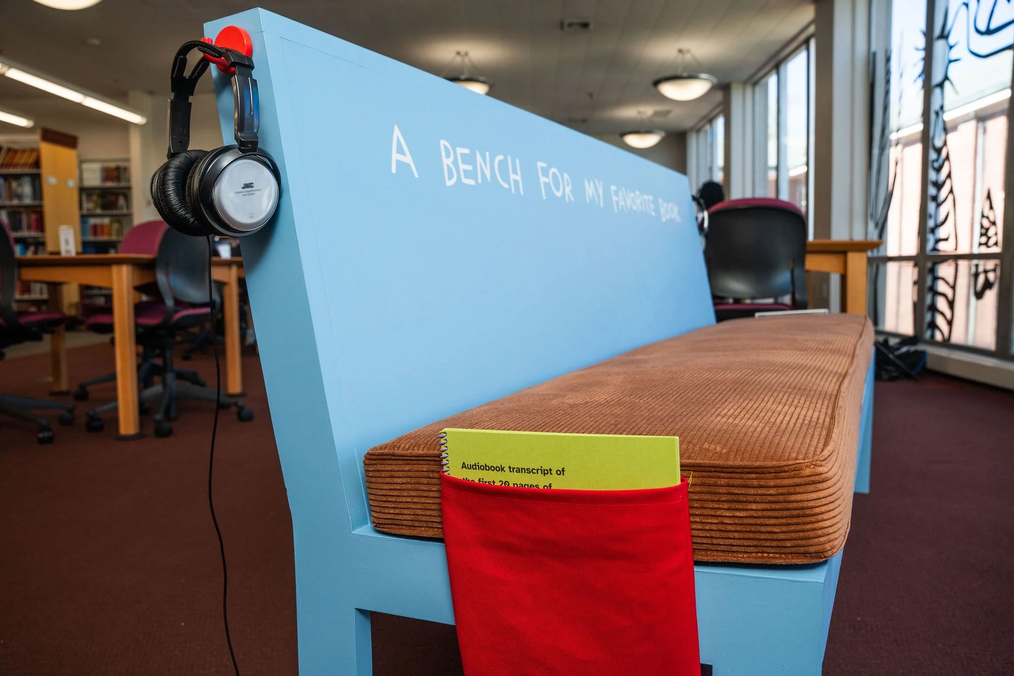 Close up of the end of this bench with a red canvas pocket for holding the audiobook transcript. A wire hangs on the floor connecting to headphones out of view.