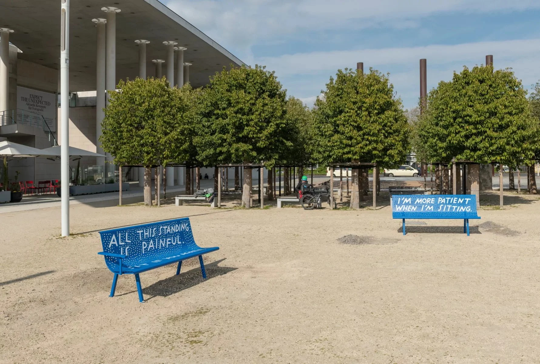 Two bright blue benches with white text sit on a dirt ground next to a line of small trees. One bench reads, "All this standing is painful." The other reads, "I'm more patient when I'm sitting." A modern building is visible in the background.