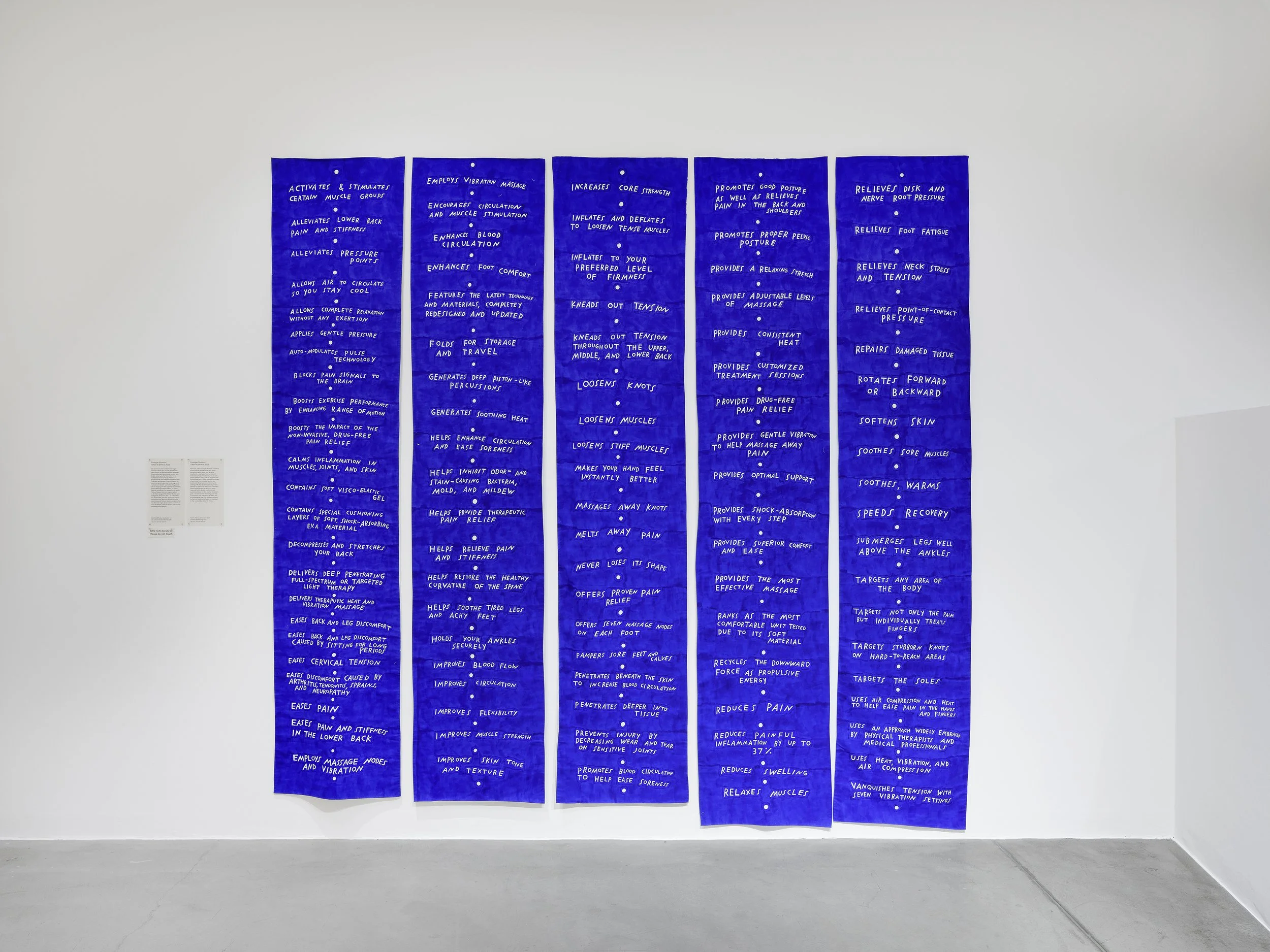 Five long blue banners hand on a wall. There is lots of small white text listed on the banners with copy from pain relief product advertising like “speeds recovery” and “soothes, warms”