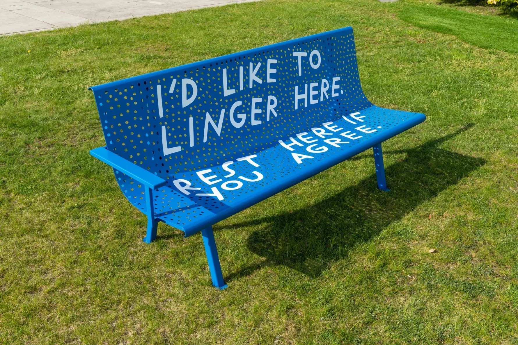 A bench install on green grass on a sunny day.  The bench is bright blue with white text that reads: “I’d like to linger here. Rest here if you agree.”