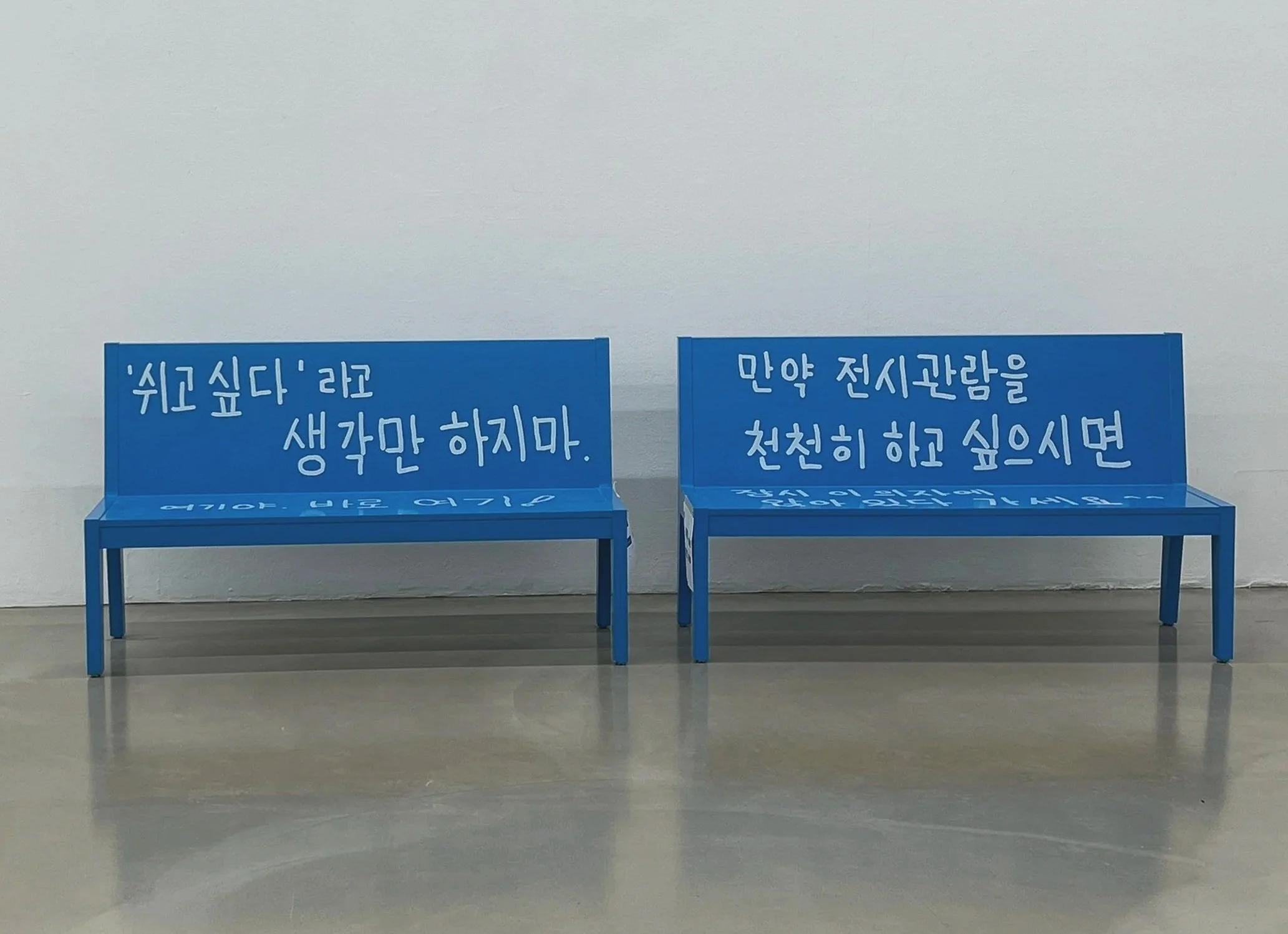 Two blue benches sit against a white wall on a polished concrete floor. Each bench has Korean text  in white handwriting.