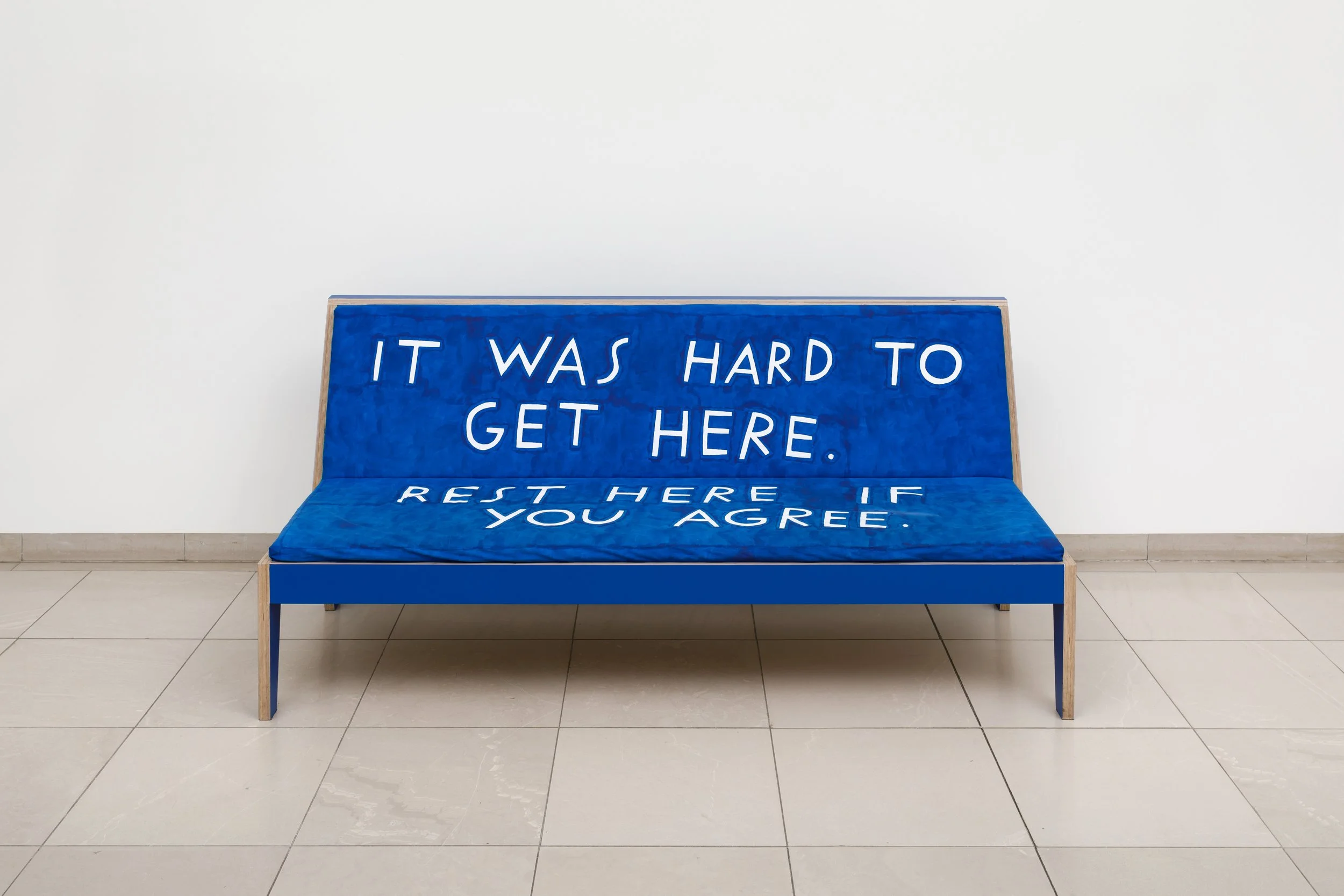 A blue padded bench with a velvet-like texture. White handwritten text reads "It Was Hard to Get Here. Rest here if you agree."