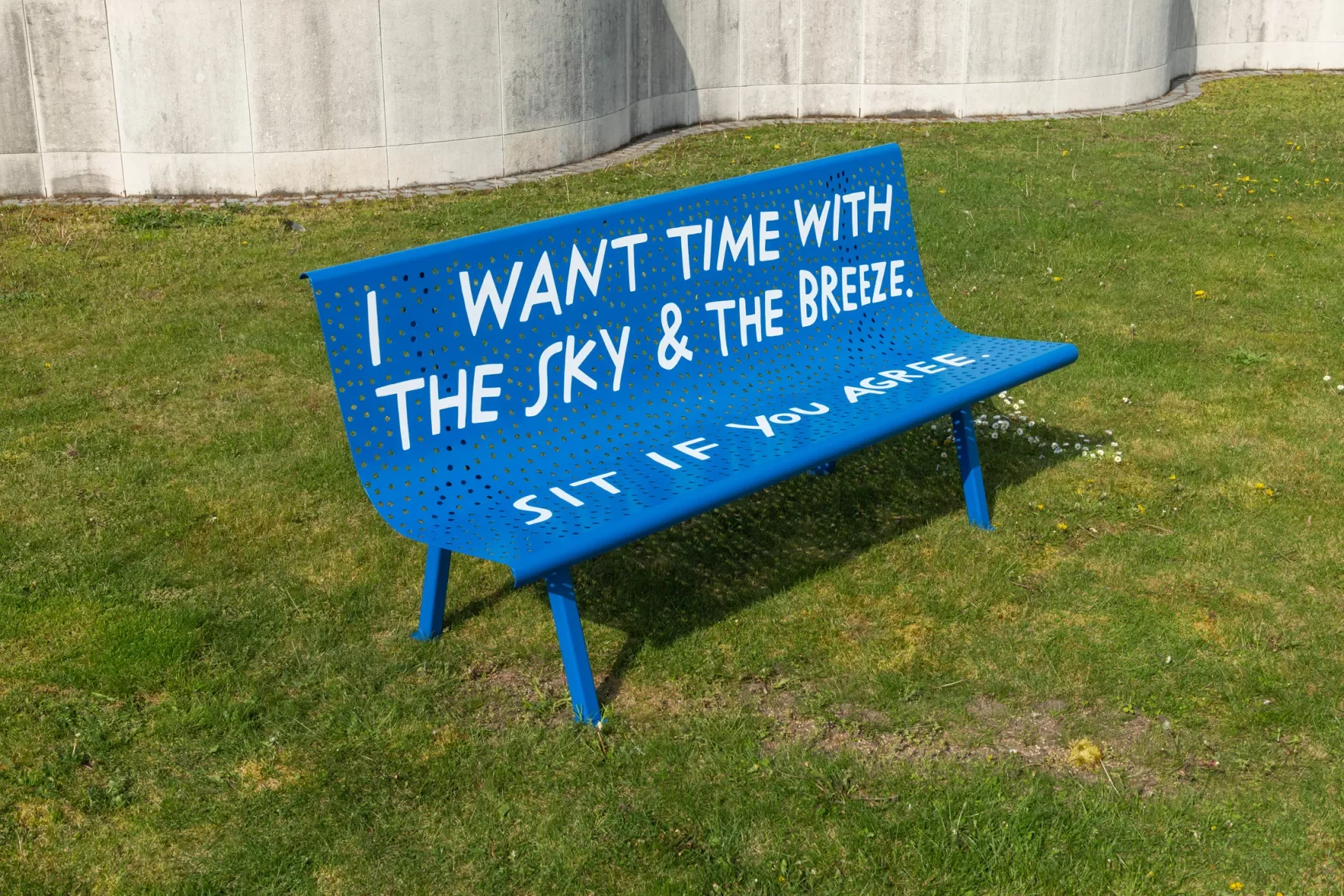 A blue metal bench sits on a green gras lawn. It has a rounded back with perforated inexact circle holes.  In white handwritten text, the bench reads “I want time with the sky & the breeze. Sit if you agree.”