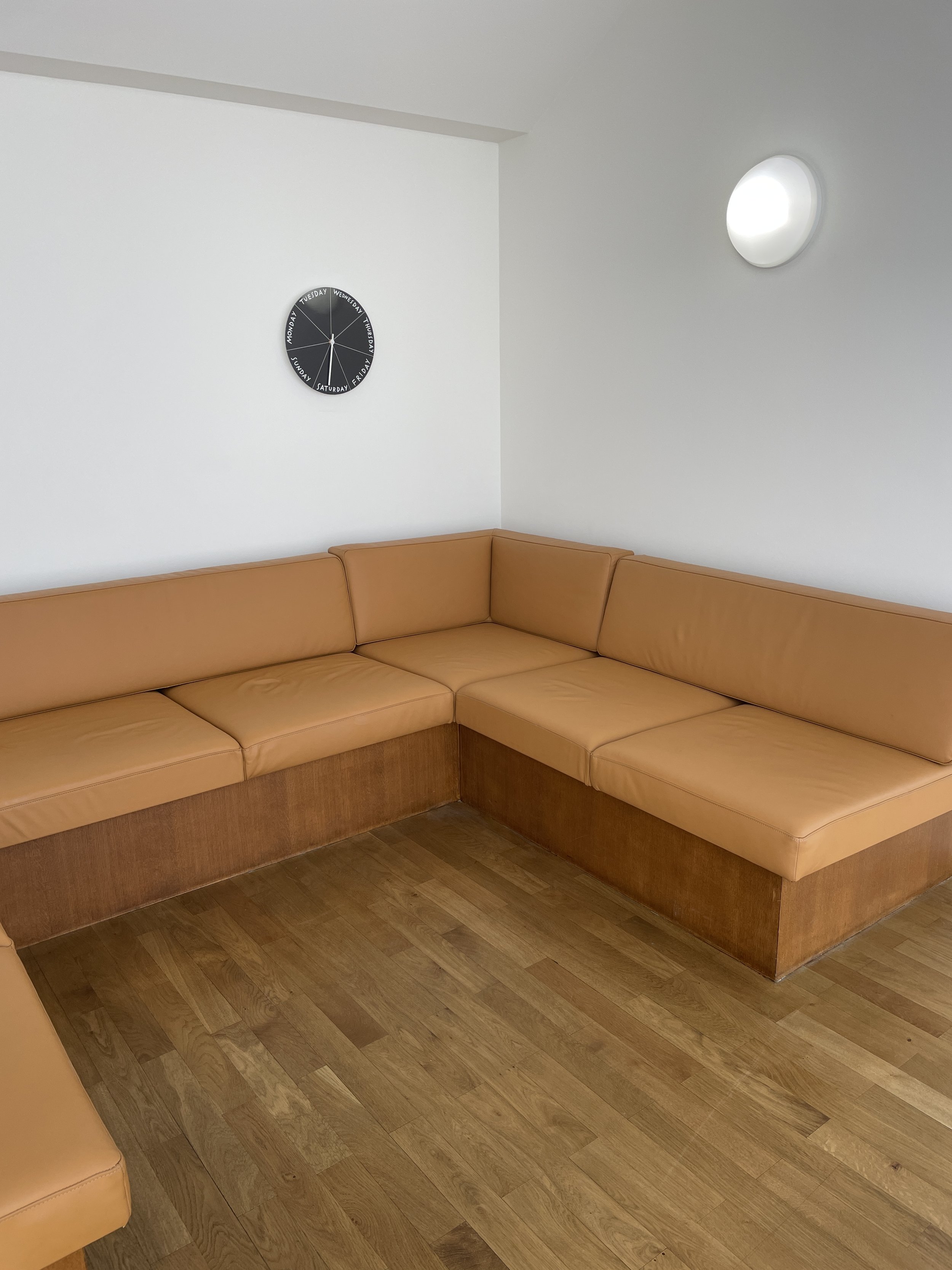 A day clock hangs above a tan, u-shaped, built-in couch.