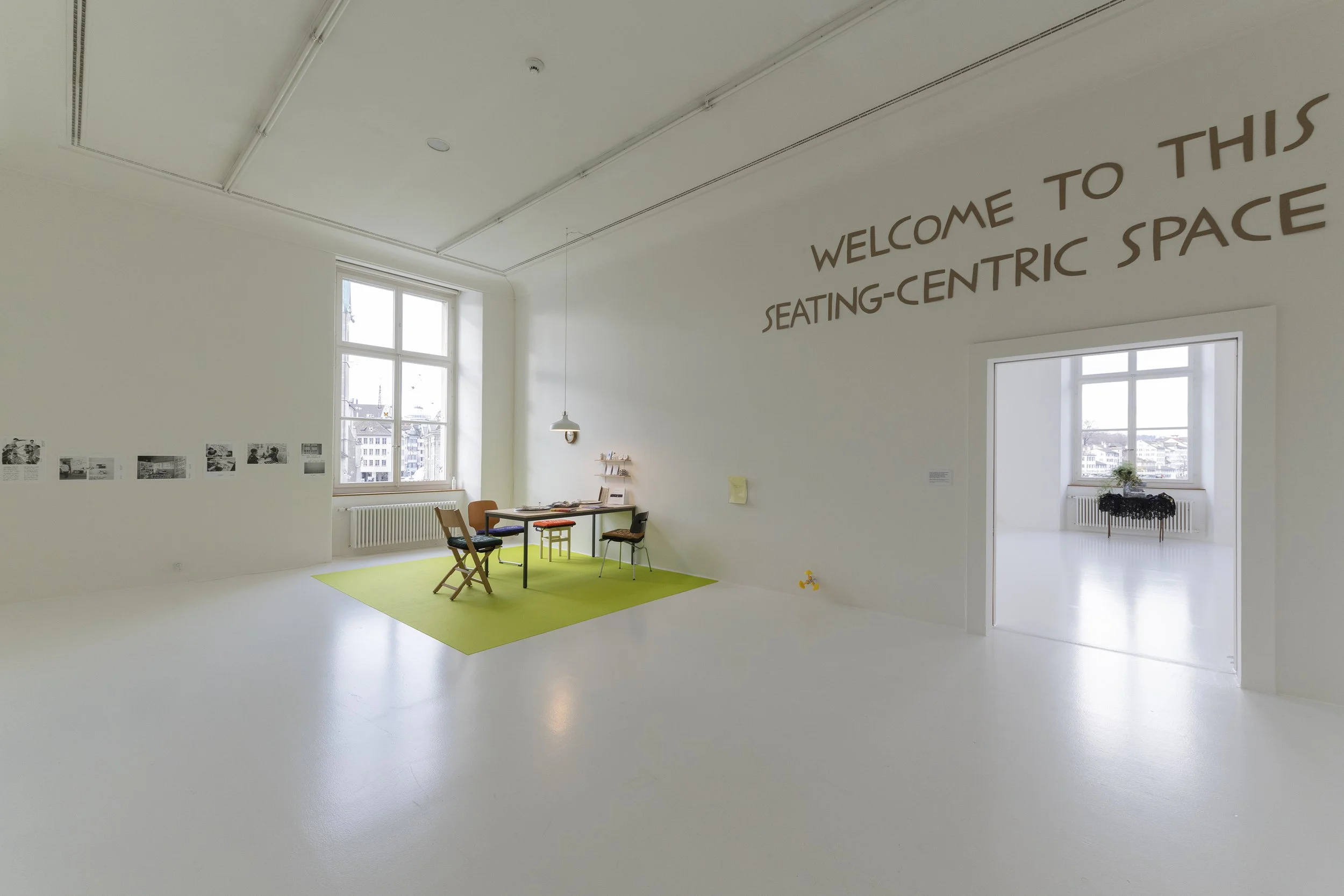 A zoomed out view showing that the table is in the corner of a large gallery space. The is a nearby window, posters on the wall, and large text that is cut off but says "welcome to this seating-centric space."