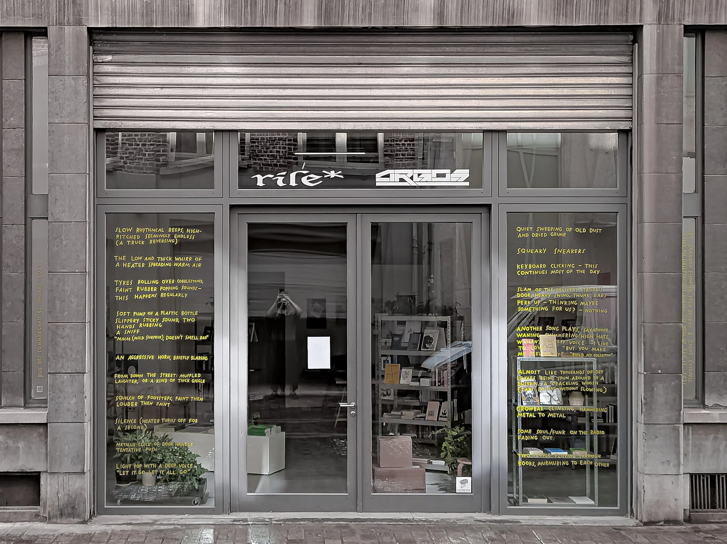 A glass storefront set in a concrete building. Flanking the glass doors are two windows filled with yellow vinyl text. Above the door it says “rile*” and “ARGOS” and through the glass, I see some bookshelves and boxes.
