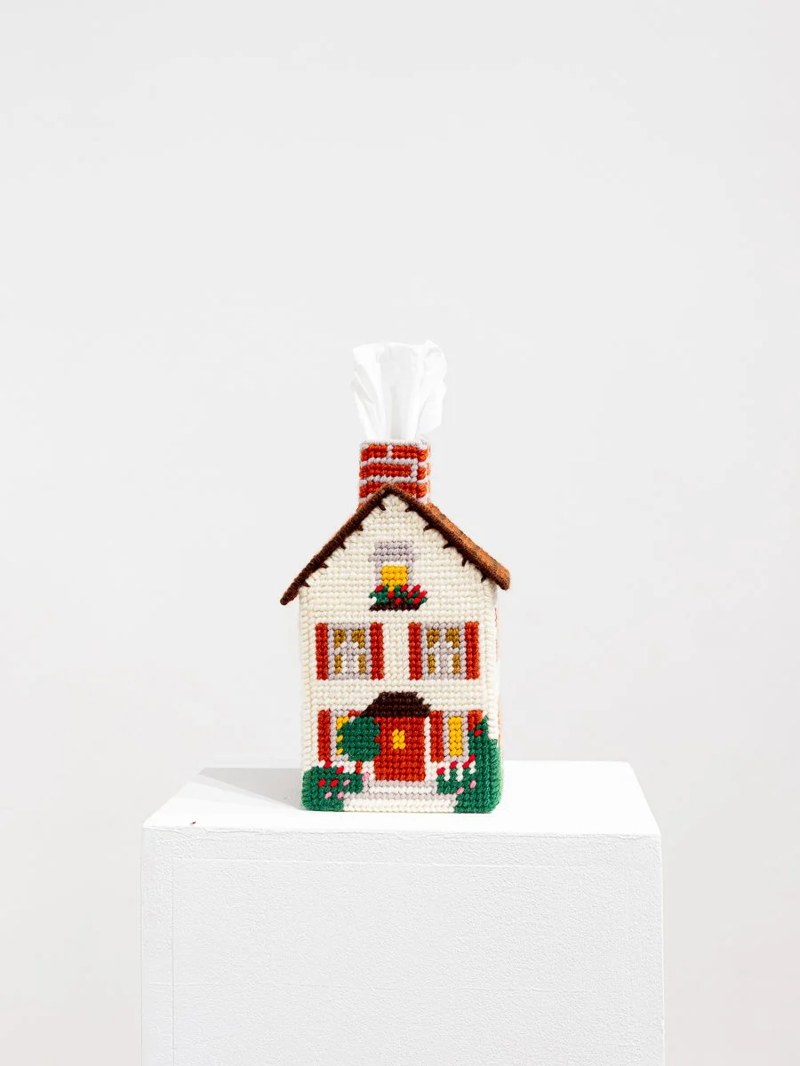 A house-shaped tissue-box cover sits on a pedestal. It’s needled pointed with doors, windows, greenery and flowers. The tissue comes out the chimney like smoke.