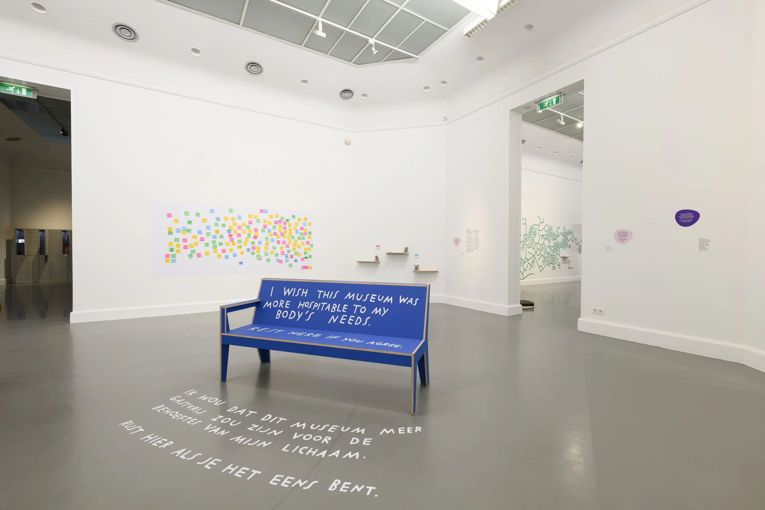 A blue bench in a gallery space with white text reading, “I wish this museum was more hospitable to my body’s needs. Rest here if you agree,”  the same message is translated in Dutch on the floor.