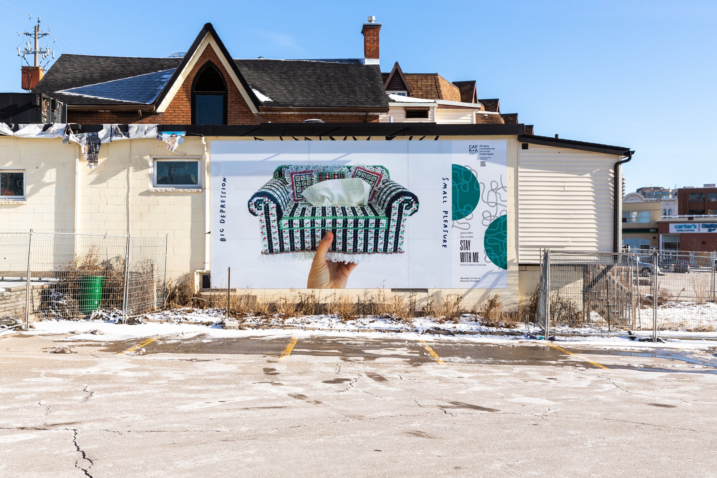 An outdoor billboard mounted on the side of a building shows a photograph of a green and white cross stitched tissue box shaped like a couch, held up by a hand. The image is framed by handwritten text, "Big Depression" and "Small Pleasure"