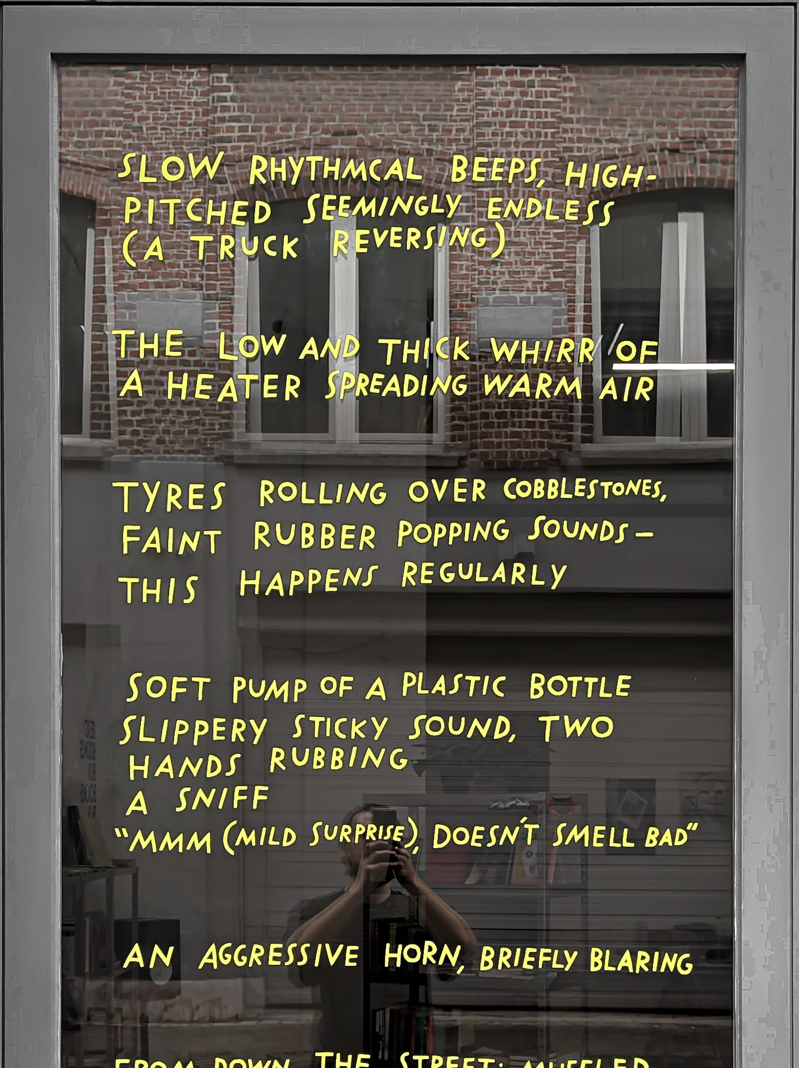 A close up of an installation of vinyl text on a dark window. The text is yellow and in handwritten in blocky, uneven capitals. A reflection of a person taking the photo with an iPhone is behind the text.