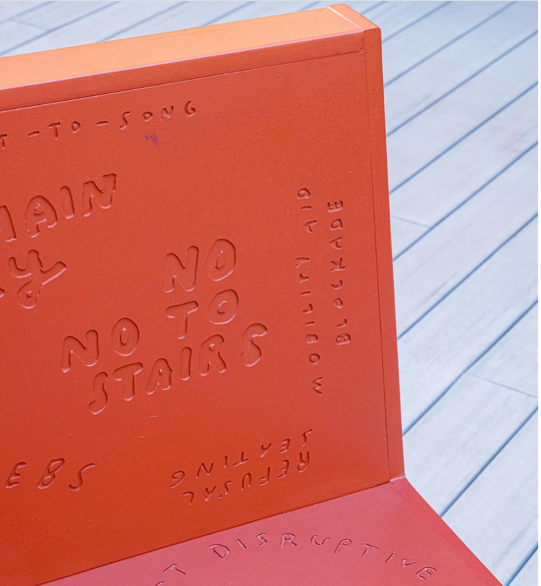 The back corner of a bench painted in an orange-red color sits on an outdoor patio floor. Words are engraved into the bench, such as “no no to stairs” and “refusal seating”.