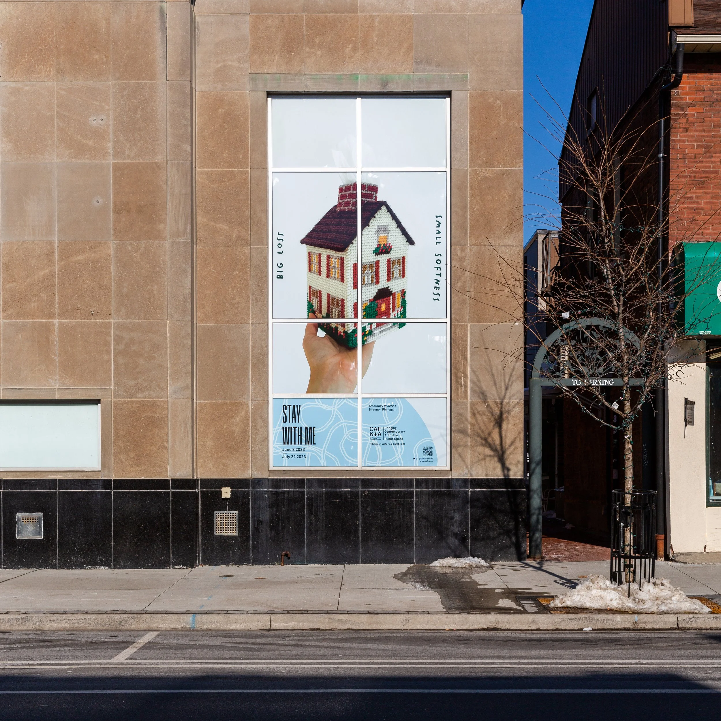 A billboard installed in a large multi-paned window on the side of a building shows a needlepointed tissue box shaped like a house, held in my hand. My handwriting reads "Big Loss" and "Small Softness" with the exhibition's branding along the bottom.