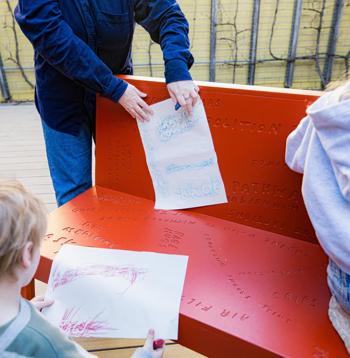 Two kids and an adult make rubbings on a red-orange bench with engraved text. The rubbings are shown on the papers held by the adult and kid.
