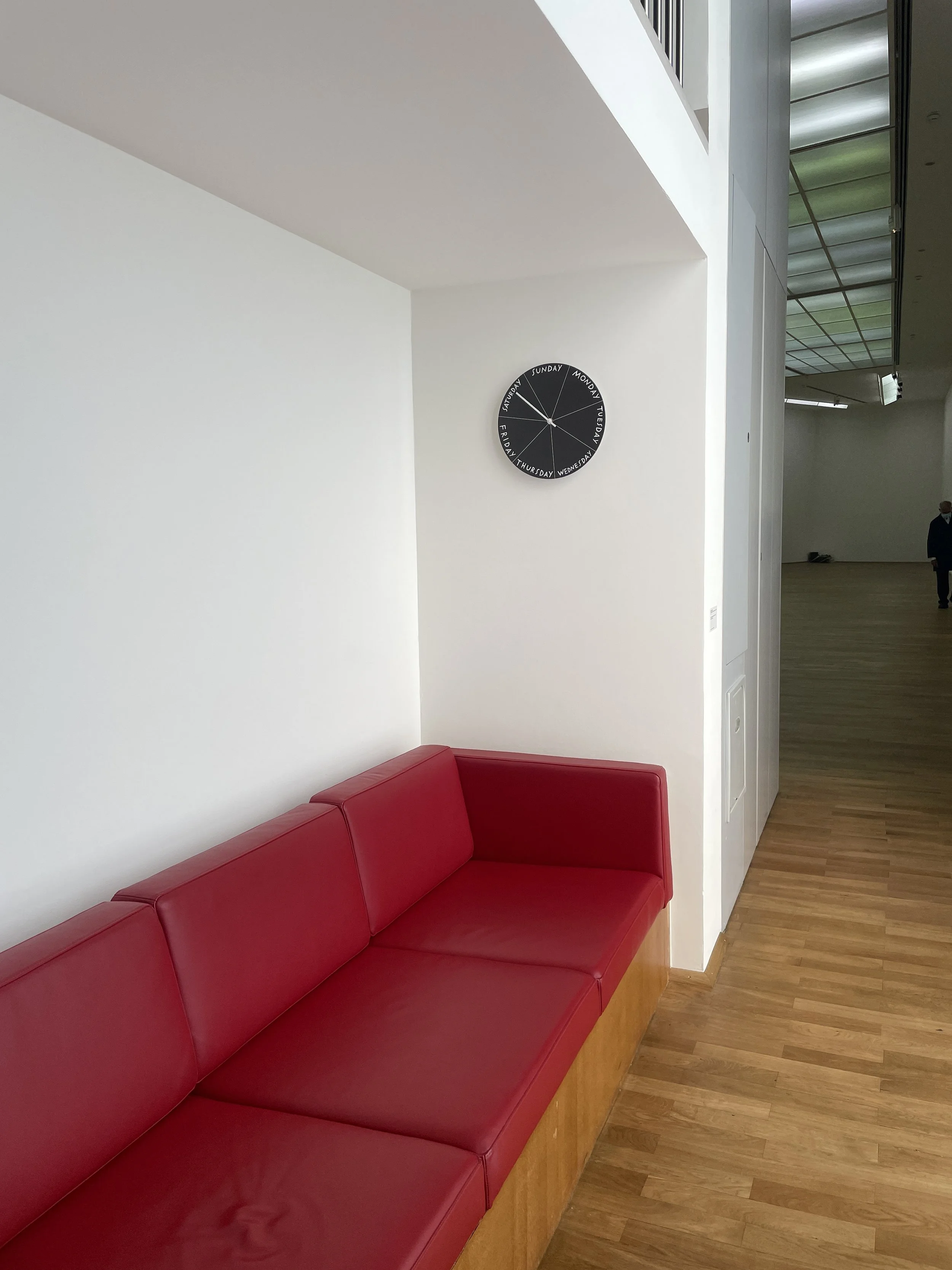 A clock hangs above a cheery, red, built-in couch. In the background, a glimpse of the exhibition space down the hall.