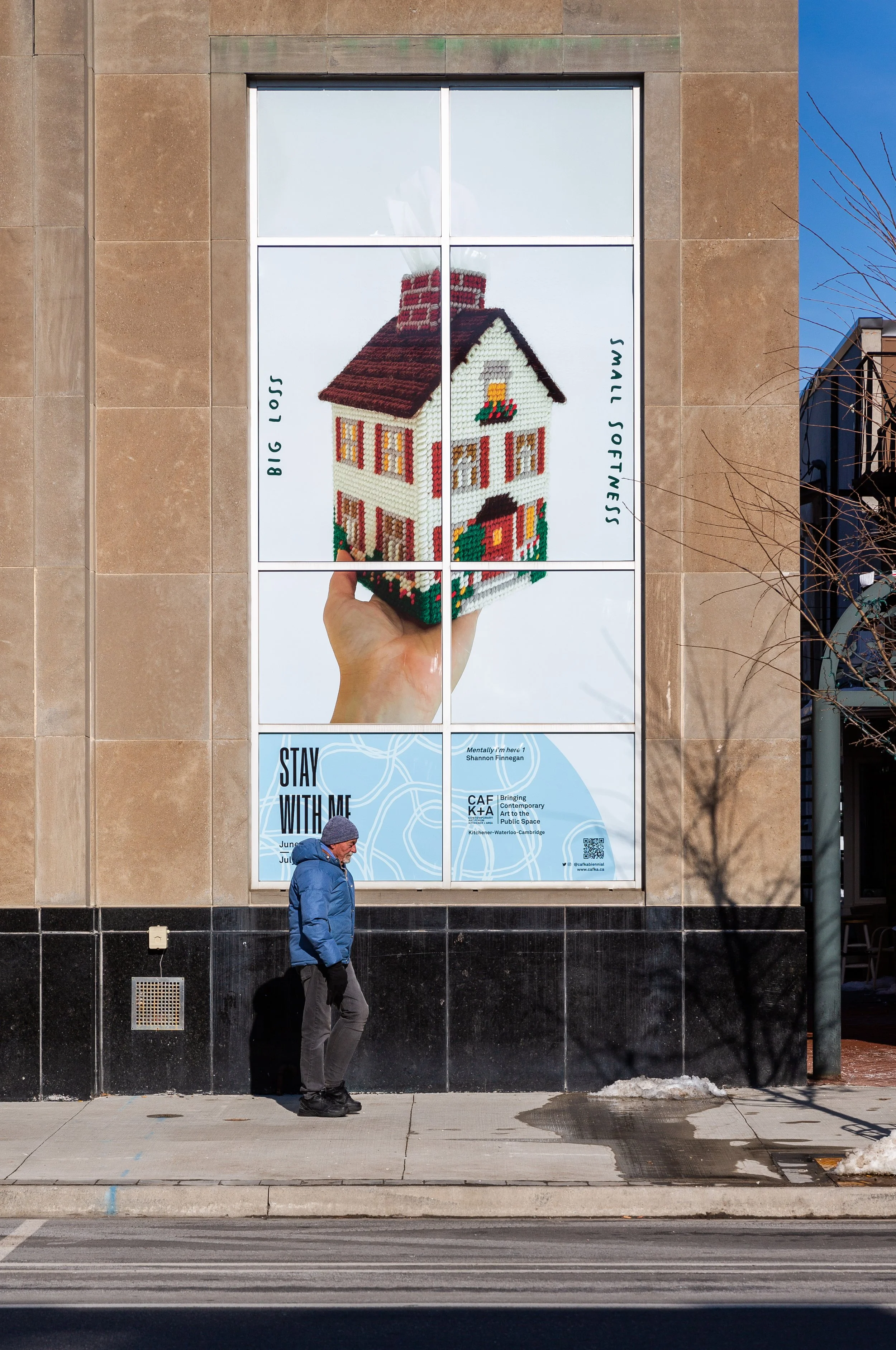 An outdoor billboard in a multi-paned window shows a cross-stitched tissue box shaped like a house, held in a hand. Text reads “Big Loss” and “Small Softness,” with CAFKA’s “Stay With Me” branding below as a passerby walks by in the foreground.
