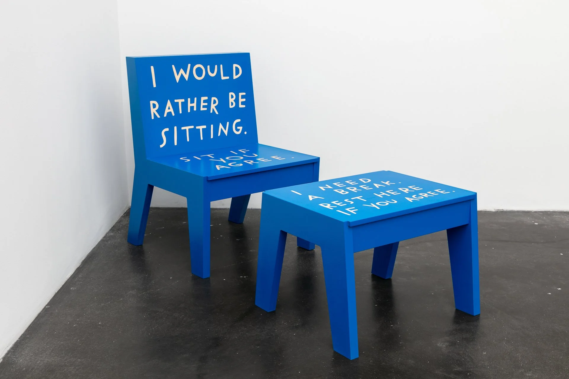A blue chair and footstool sit in the corner of a white room. The chair has text in white handwritten letters, “I would rather be sitting. Sit if you agree.” The footstool text in the same style, “I need a break. Rest if you agree.”