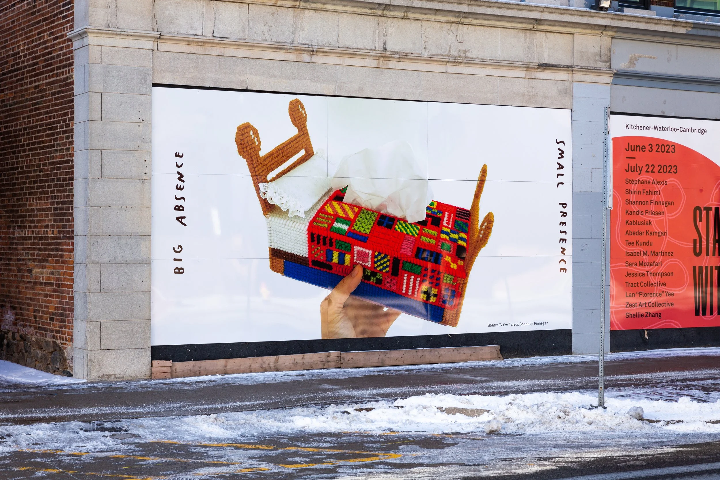 Installation view of a large panel in front of a building showing a hand holding a colorful crossed-stitched tissue box shaped like a bed. Handwritten text bookends the image, reading "Big Absence, Small Presence"