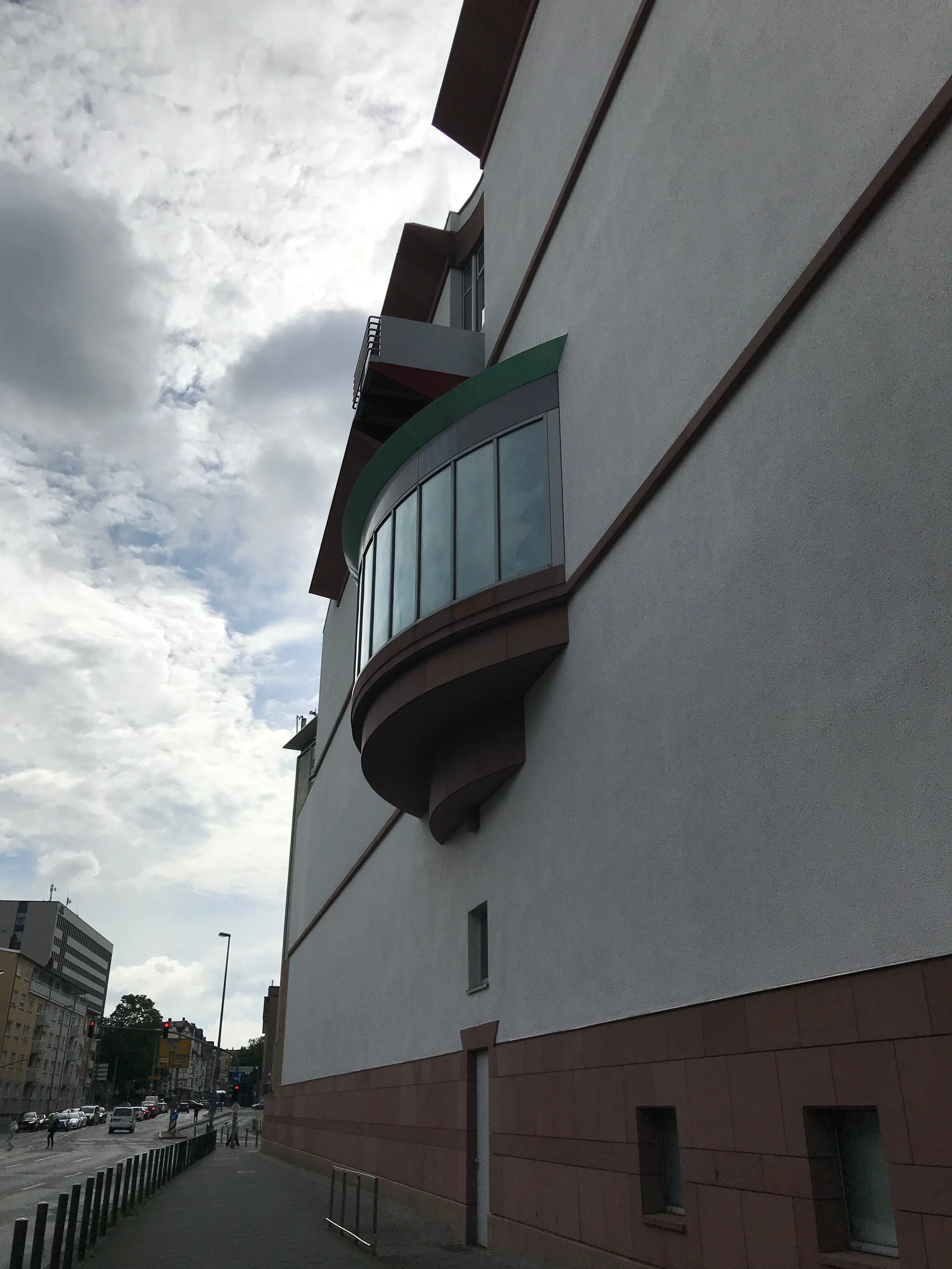 A curved glass window protrudes from the side of a large grey building above a mostly empty sidewalk and street corner. The sky is filled with bright white clouds.