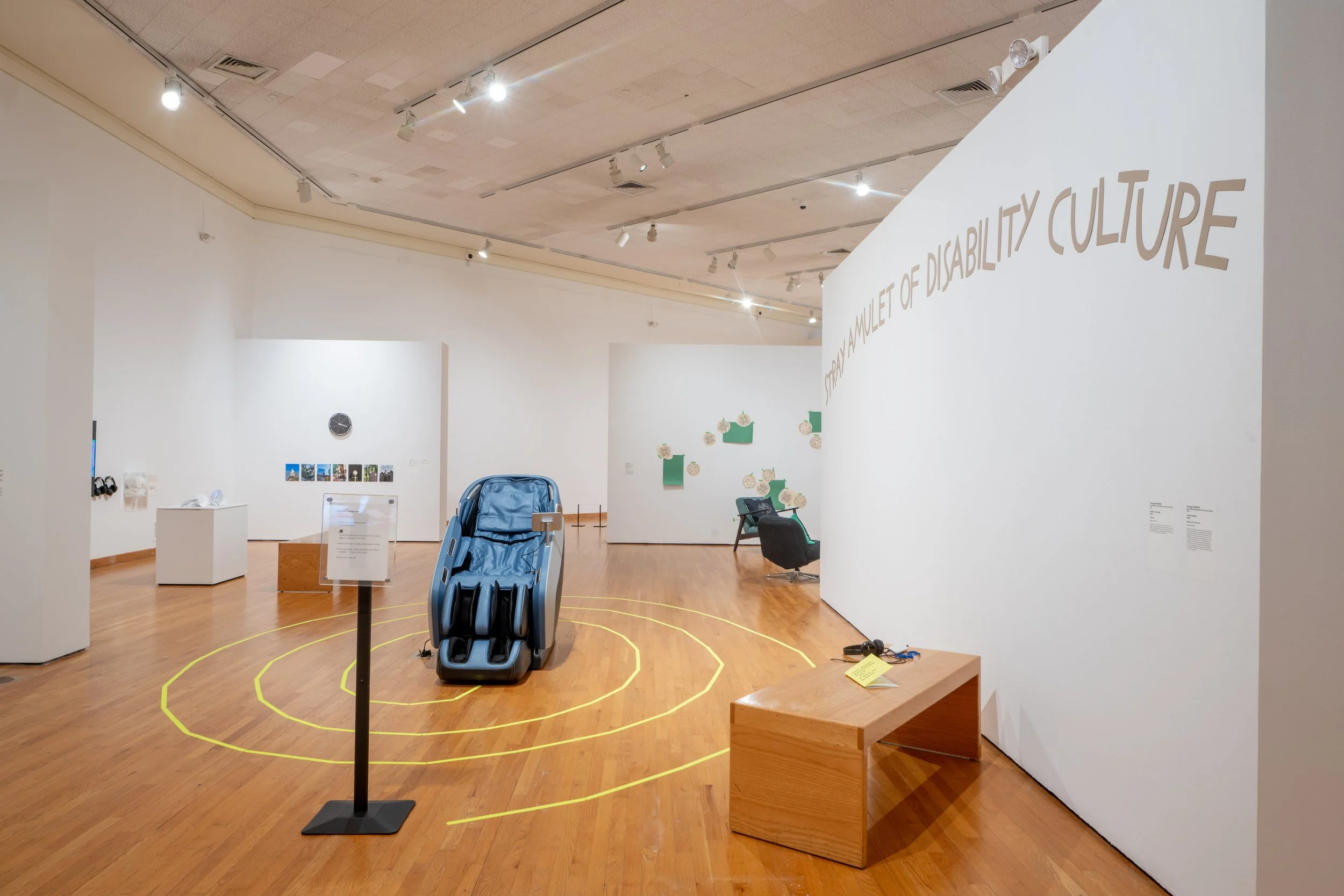 A massage chair sits within a yellow spiral on a museum gallery floor. Audio and a zine are paired with it, featuring a text composed from pain relief product ads using phrases like "eases aches and pains," and "long-lasting relief"