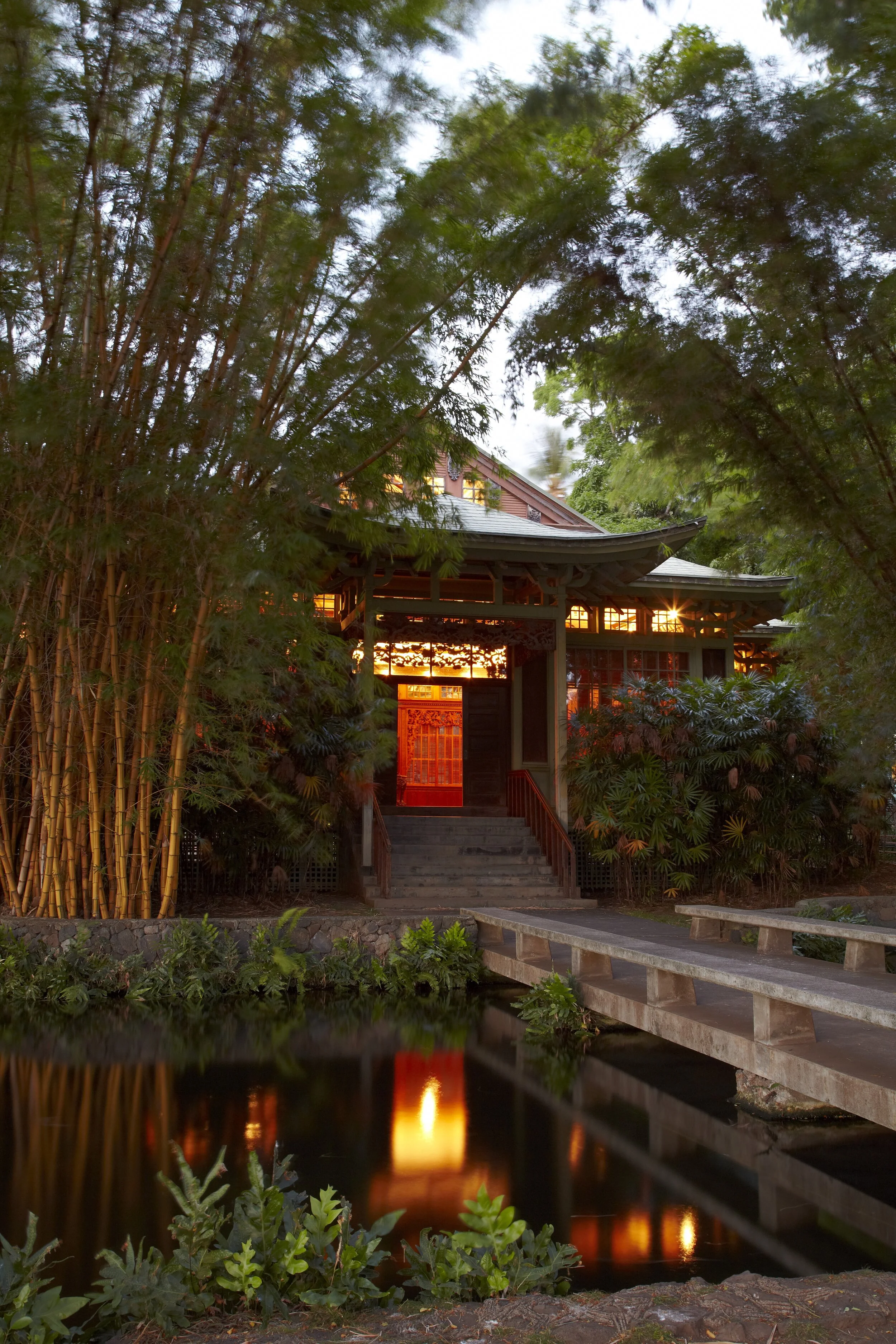 A traditional Japanese building is situated amidst dense green trees, with warm lighting emanating from inside. The structure is accessed by a small staircase and is reflected in the calm water at the front, which is bordered by lush plants.