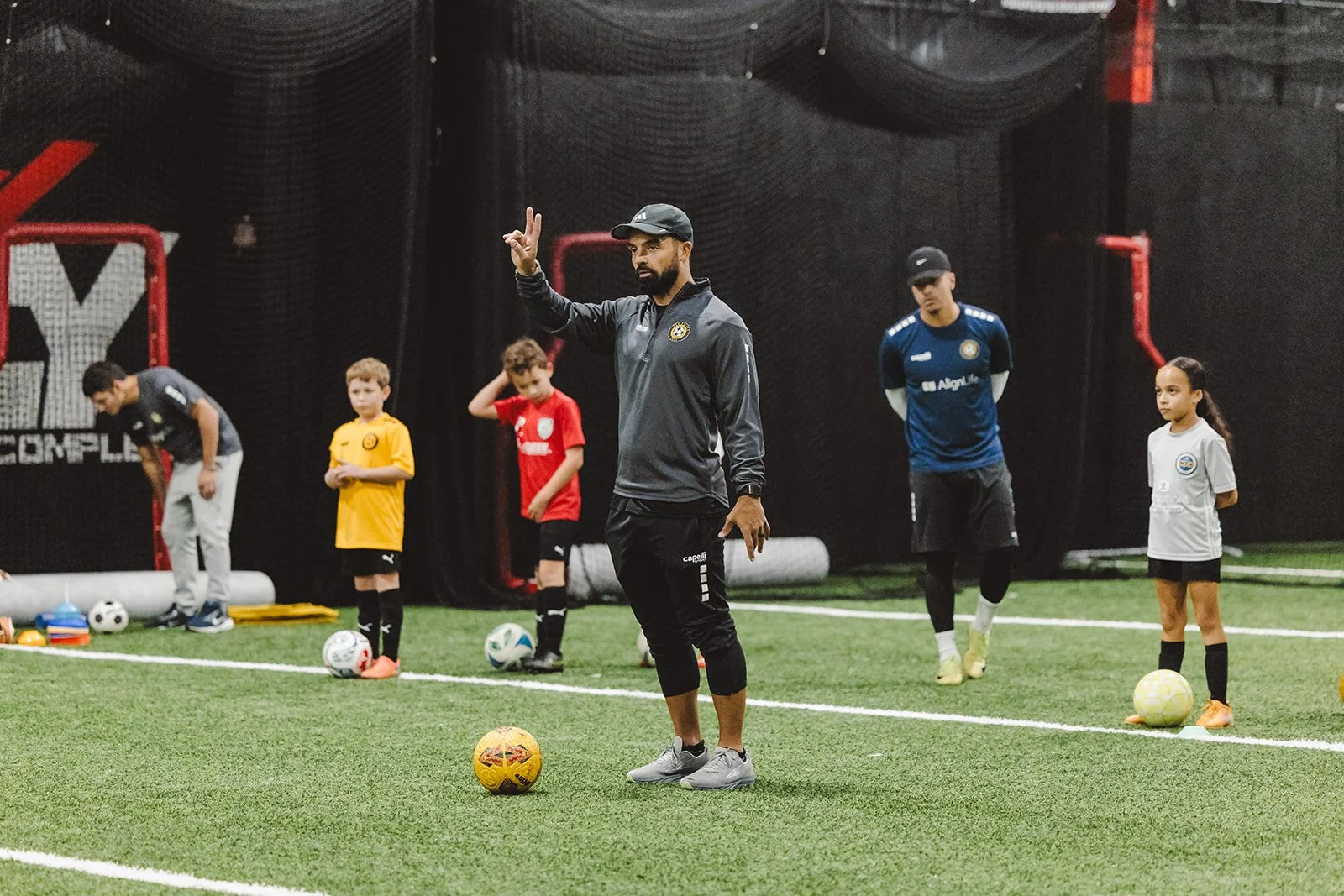 Soccer coach instructing young players in an indoor training facility, standing on artificial turf with soccer balls. The children are dressed in colorful jerseys, and the coach is making a gesture with his hand.