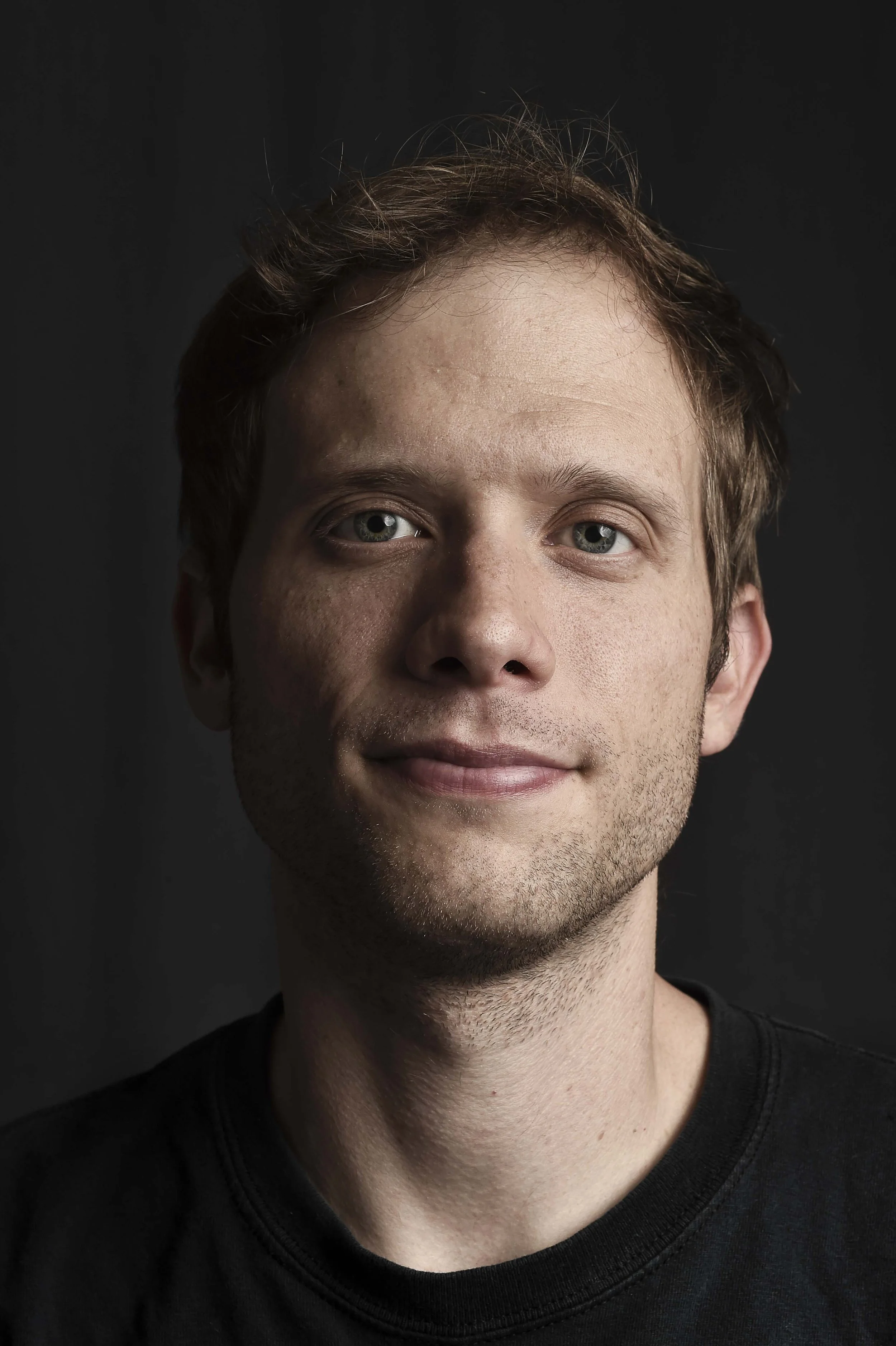 A young man with short brown hair and light skin, wearing a black shirt, looking directly at the camera with a slight smile, against a dark background.